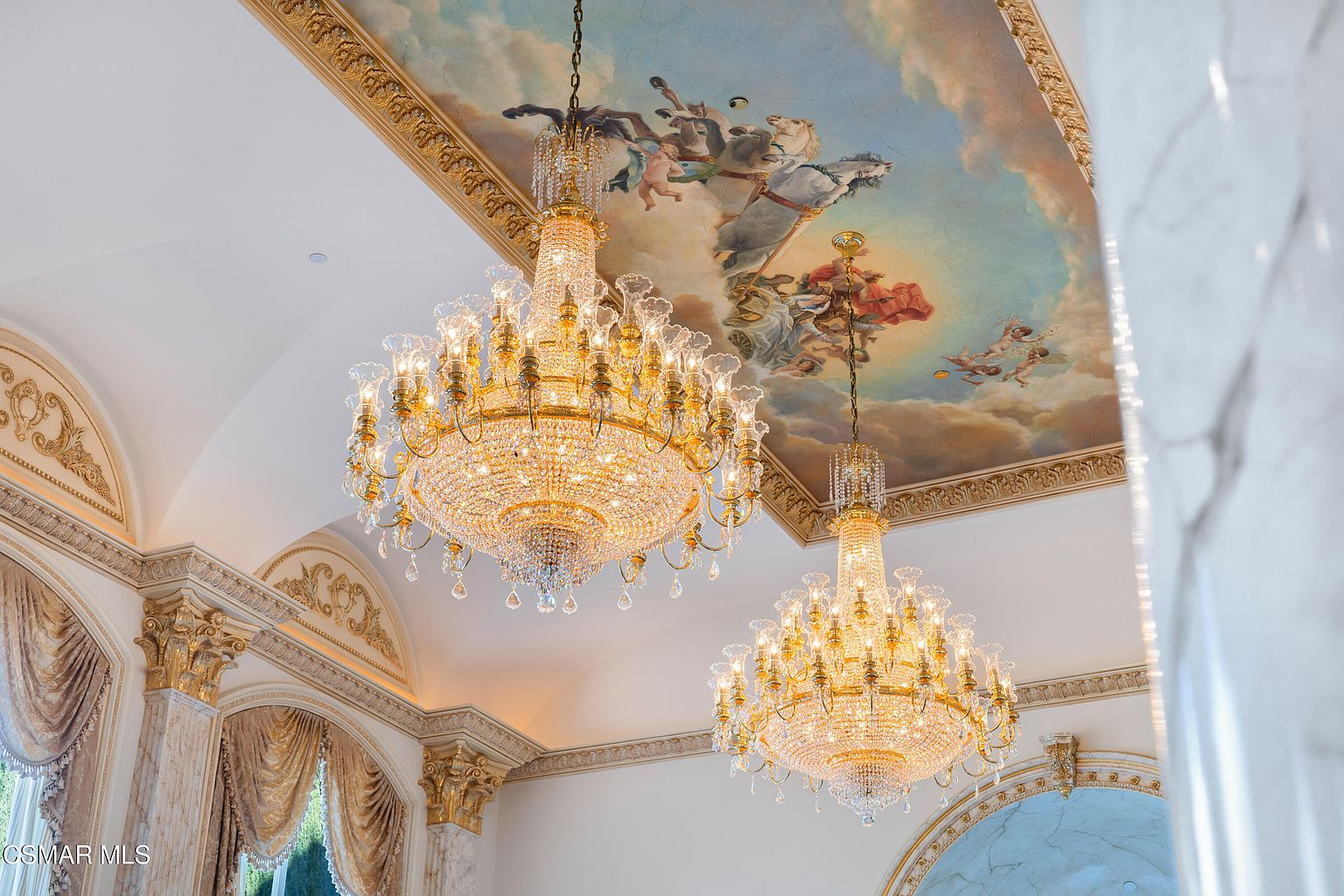 This image captures a grand, opulent ceiling featuring a detailed classical mural of figures and horses set against a sky-blue backdrop. Two massive, ornate crystal chandeliers hang prominently from the ceiling, serving as the focal point of the room's luxurious design. The surrounding architecture includes intricate gold-leaf molding and marble-like wall textures, evoking a sense of regal, palatial elegance.