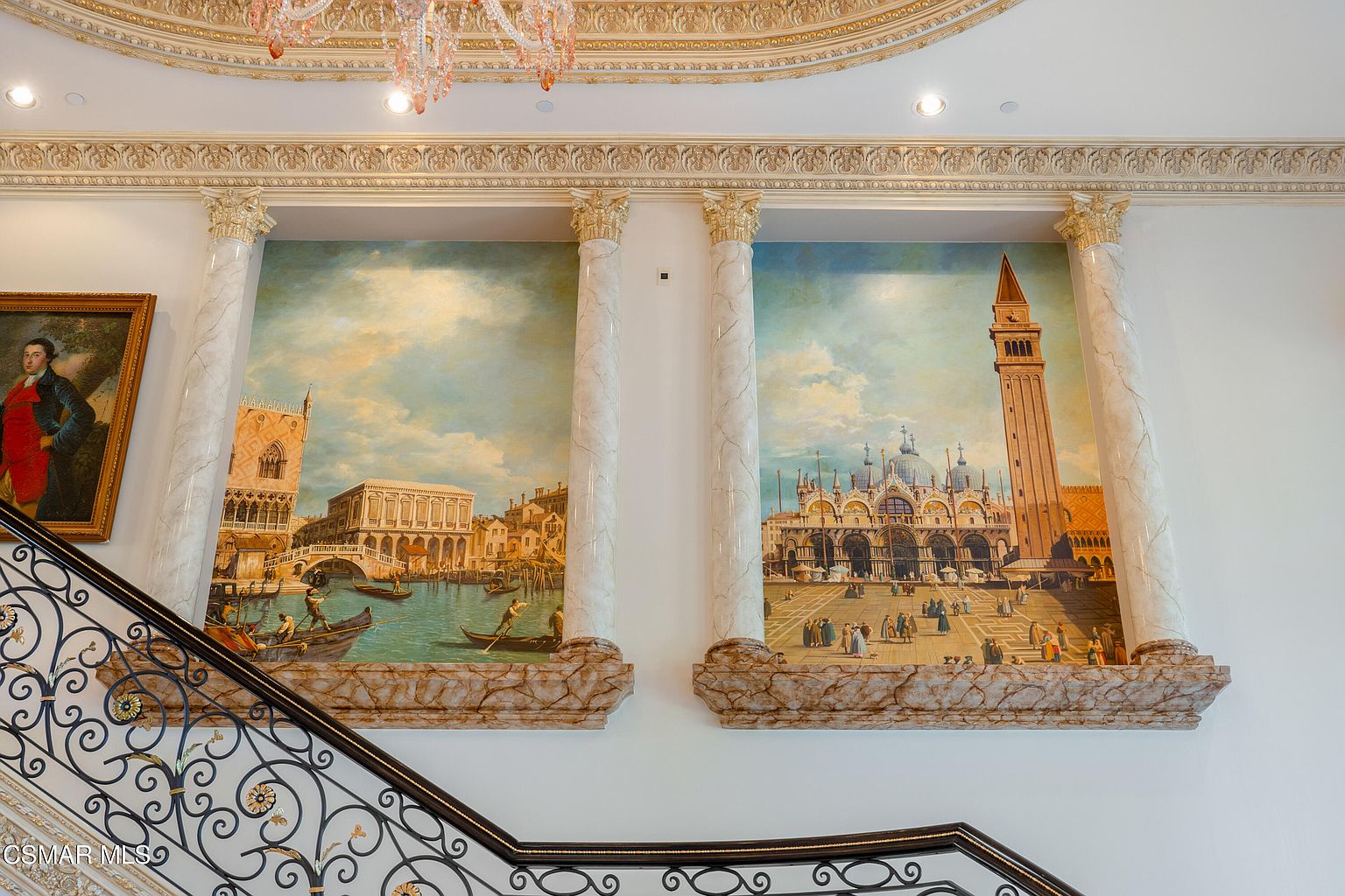 This grand interior space features a sweeping staircase with an ornate wrought-iron railing leading up to a wall adorned with two large, classical-style murals depicting Venetian scenes. The area is framed by elegant marble-look columns with gold-leaf capitals, set against a backdrop of intricate crown molding and a luxurious crystal chandelier. The overall impression is one of opulent, old-world European sophistication and architectural grandeur.