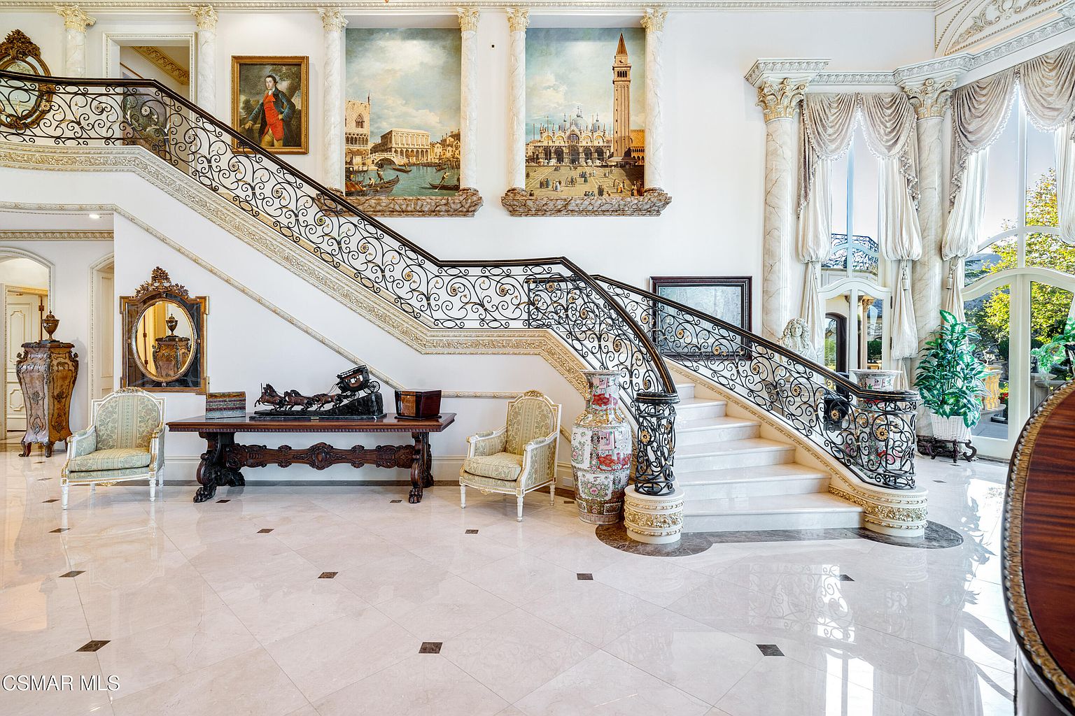 This grand foyer features a sweeping, ornate wrought-iron staircase that serves as the focal point of the opulent, high-ceilinged space. The area is decorated with classical paintings, marble flooring, and elegant furniture, creating a sophisticated and luxurious atmosphere. The perspective is a wide, eye-level shot that captures the symmetry and architectural grandeur of the entryway.