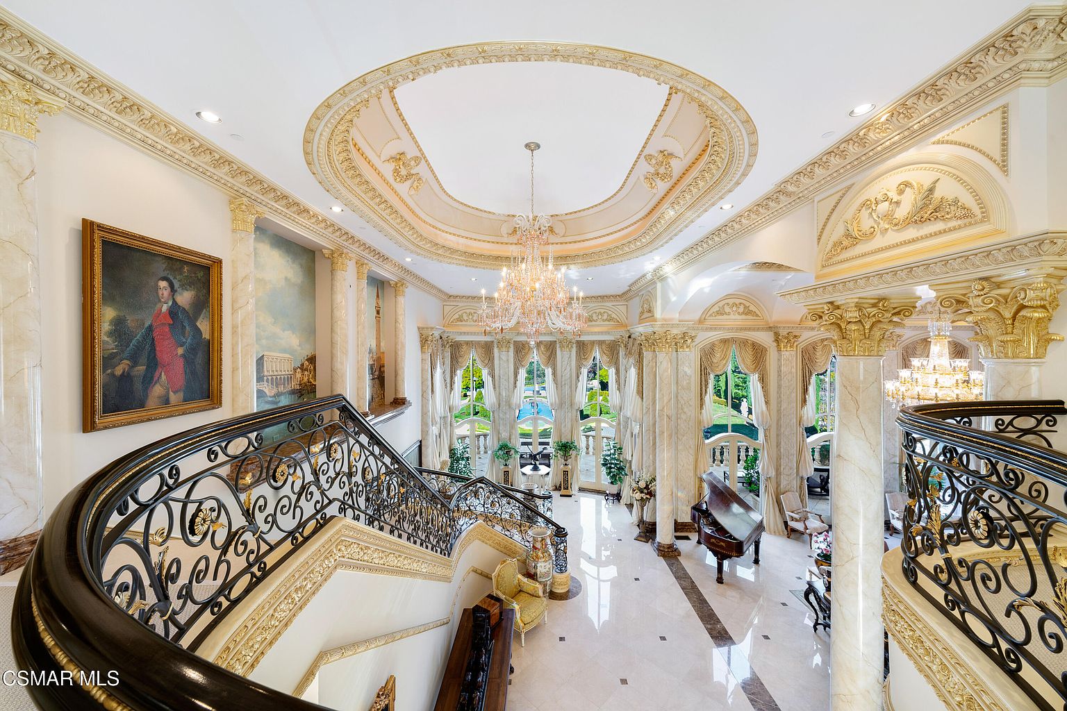 This grand foyer features a sweeping, ornate wrought-iron staircase that descends into a palatial living space characterized by high ceilings, marble flooring, and intricate gold-leaf detailing. The room is illuminated by a magnificent crystal chandelier hanging from a recessed, decorative dome, while large arched windows provide a view of the lush outdoor landscape. The space exudes opulence and classical elegance, highlighted by a grand piano and a large portrait on the wall.