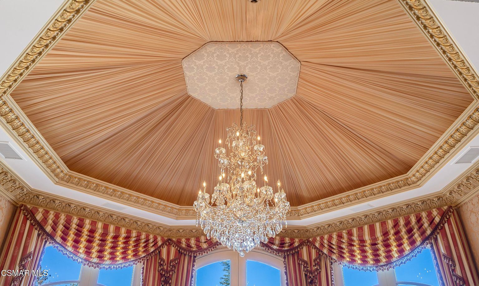 This image captures an opulent, octagonal ceiling feature characterized by draped, pleated fabric in a warm peach tone that radiates toward a central, patterned medallion. A grand, multi-tiered crystal chandelier hangs from the center, serving as the focal point of the room's sophisticated and ornate design. Below, elegant red and gold striped drapery frames the windows, contributing to a luxurious, classical aesthetic.