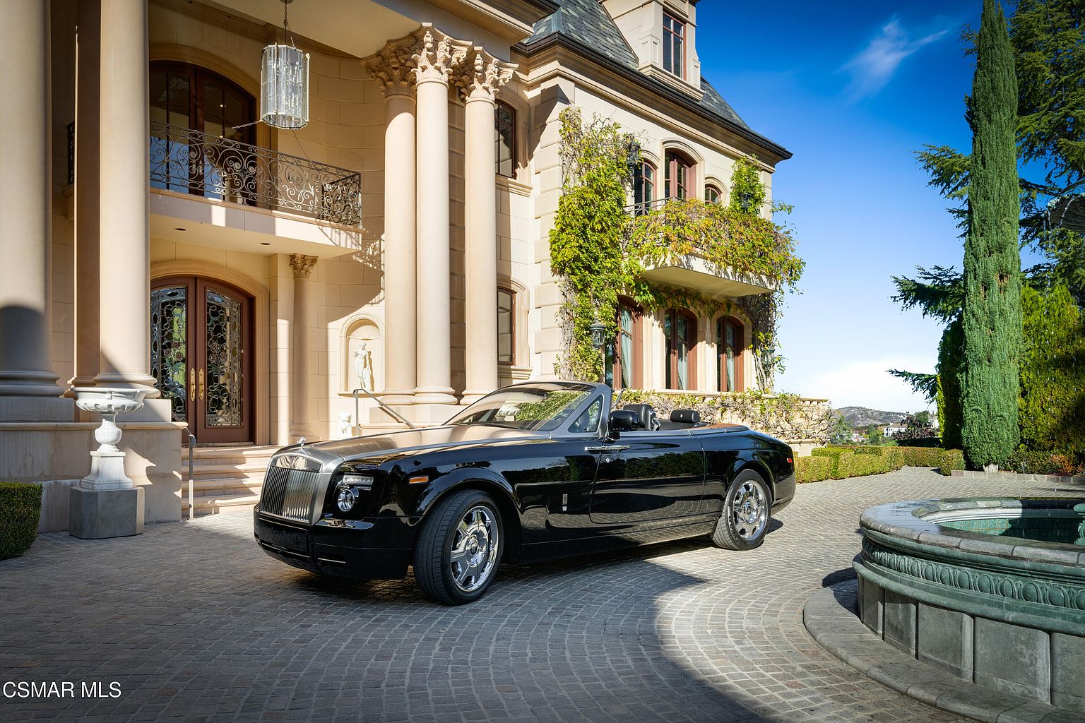 This grand estate features a sophisticated Mediterranean-inspired architectural style, highlighted by towering Corinthian columns and an ornate, arched entryway. A luxurious black Rolls-Royce convertible is parked prominently on the circular cobblestone driveway, which is accented by a stone fountain in the foreground. The scene exudes opulence and prestige, framed by lush greenery and a clear blue sky.