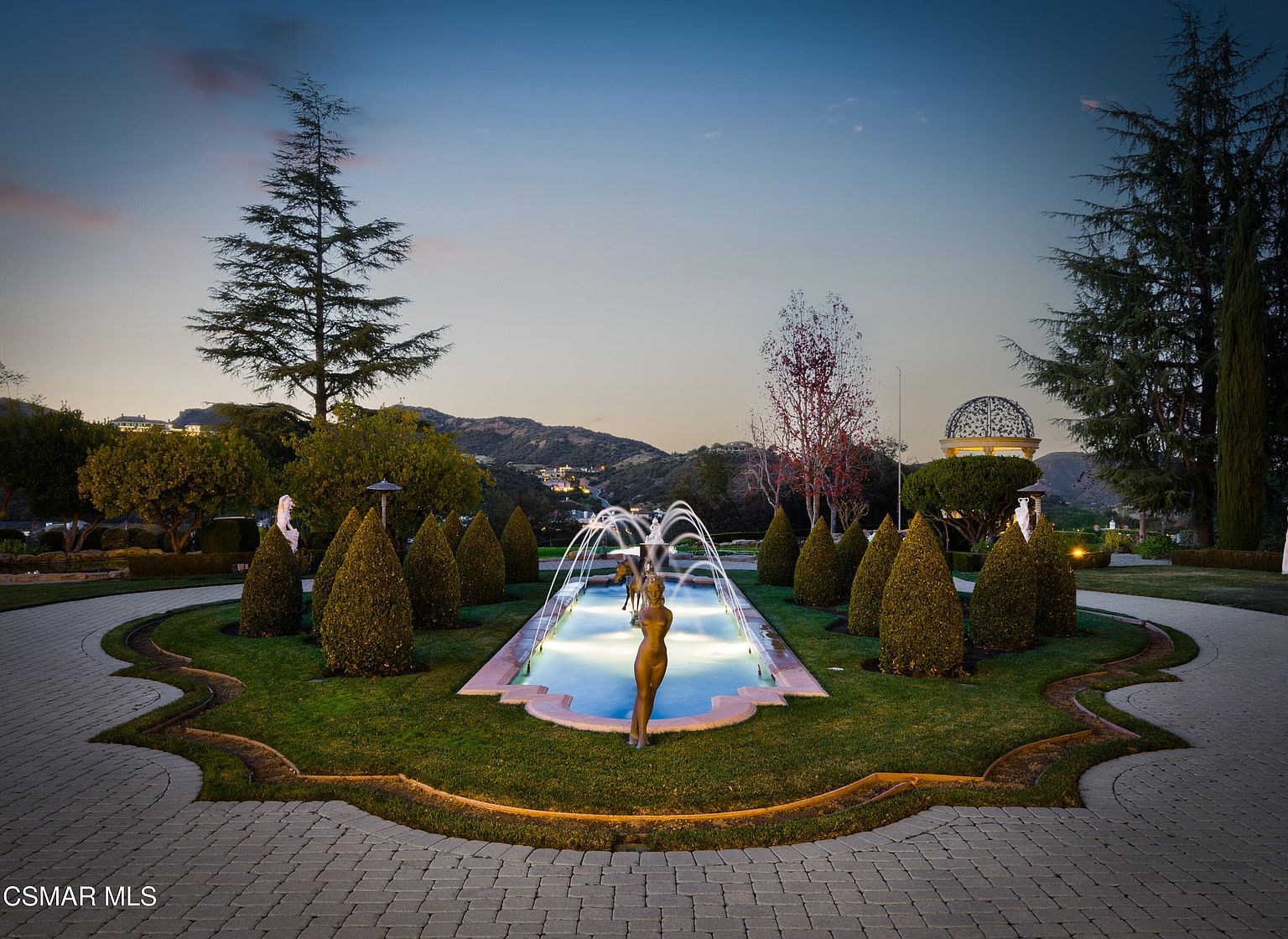 This elegant garden scene features a symmetrical, illuminated water fountain as the central focal point, surrounded by manicured conical hedges and lush green lawn. The perspective is a wide, eye-level shot looking toward a scenic mountain backdrop at dusk, creating a sophisticated and serene atmosphere. The paved circular driveway and classic statue accents enhance the luxurious, estate-like feel of the property.
