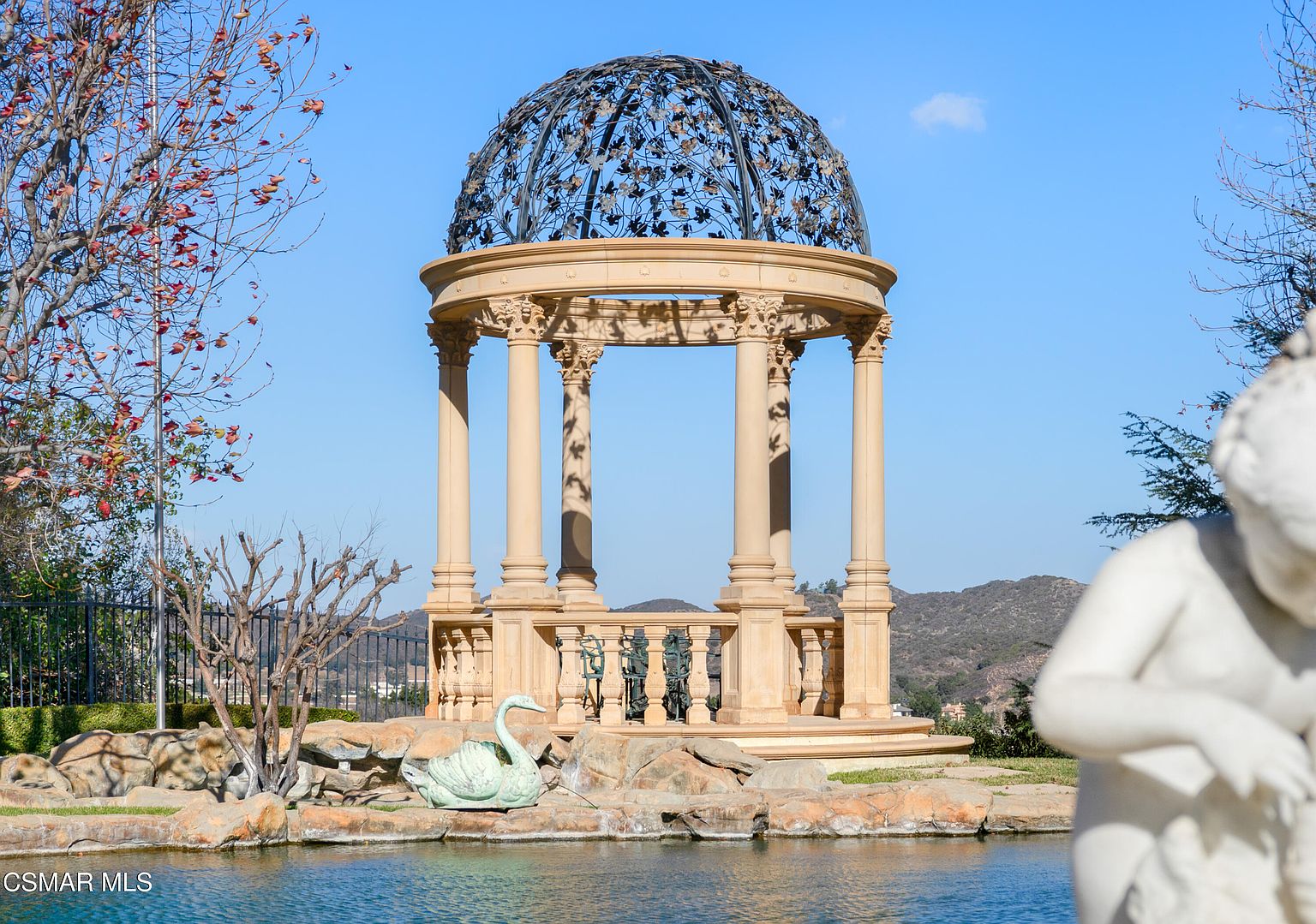 This elegant garden scene features a classical-style stone gazebo with an intricate metal dome, set against a backdrop of rolling hills and a clear blue sky. In the foreground, a serene pond is accented by a decorative swan sculpture and a partial view of a white marble statue, creating a sophisticated and tranquil atmosphere. The composition highlights the luxurious landscaping and architectural detail, perfect for showcasing a high-end estate property.