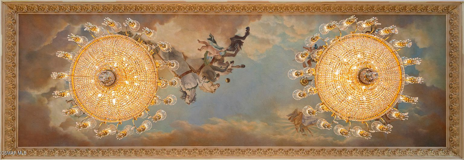 This image captures a grand, ornate ceiling featuring a classical mural of figures in a sky-like setting, framed by an intricate, gilded molding. Two large, multi-tiered crystal chandeliers hang prominently, casting a warm, brilliant glow across the painted scene. The perspective is a direct upward view, emphasizing the opulent and artistic craftsmanship of the ceiling design.