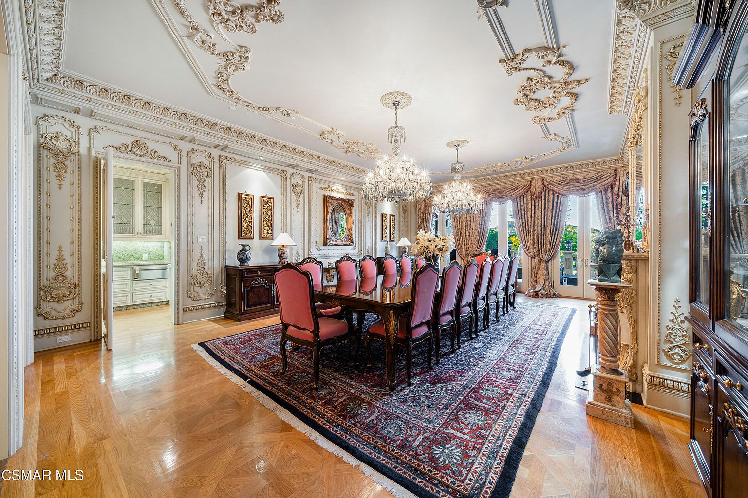 This opulent formal dining room features intricate crown molding, ornate wall paneling, and two grand crystal chandeliers that serve as the focal point. A long, polished wooden table is surrounded by elegant pink-upholstered chairs, all set atop a large, traditional patterned area rug. The room is framed by floor-to-ceiling drapery and offers a glimpse into an adjacent butler's pantry, creating a sophisticated and luxurious atmosphere.