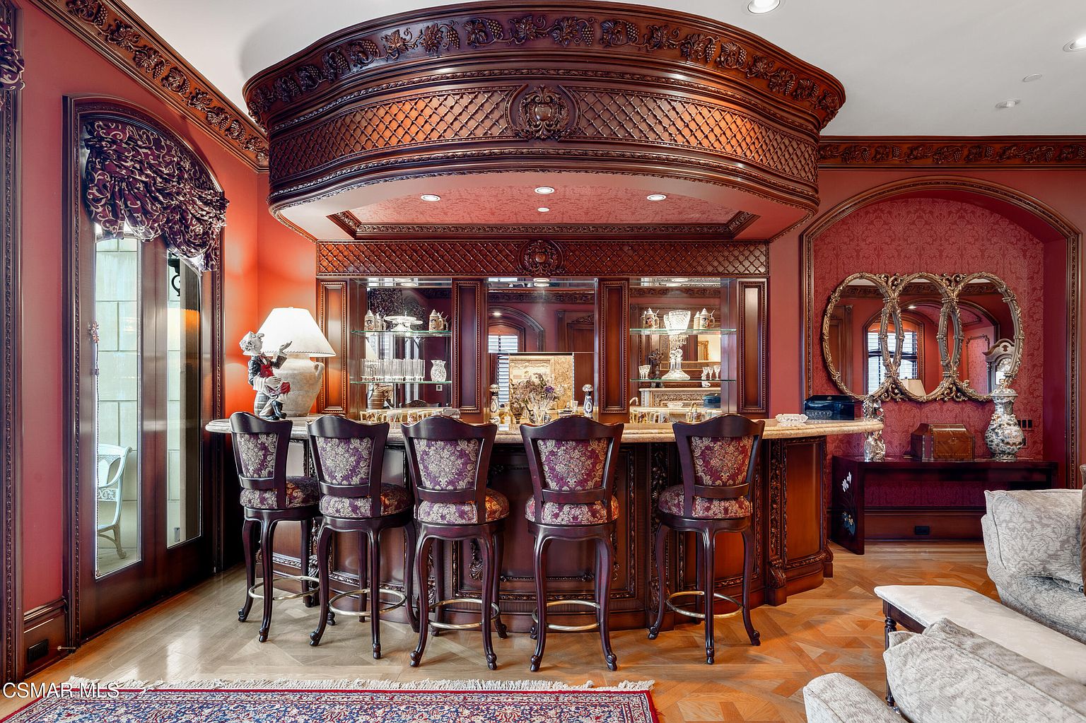 This opulent home bar area features a grand, custom-built wooden bar with intricate carvings, a lattice-patterned canopy, and a polished stone countertop. Five ornate, upholstered bar stools are arranged in a semi-circle, set against a backdrop of rich red walls, mirrored shelving, and a decorative gold-framed mirror. The space exudes a sophisticated, classic luxury aesthetic, perfect for entertaining in a formal residence.