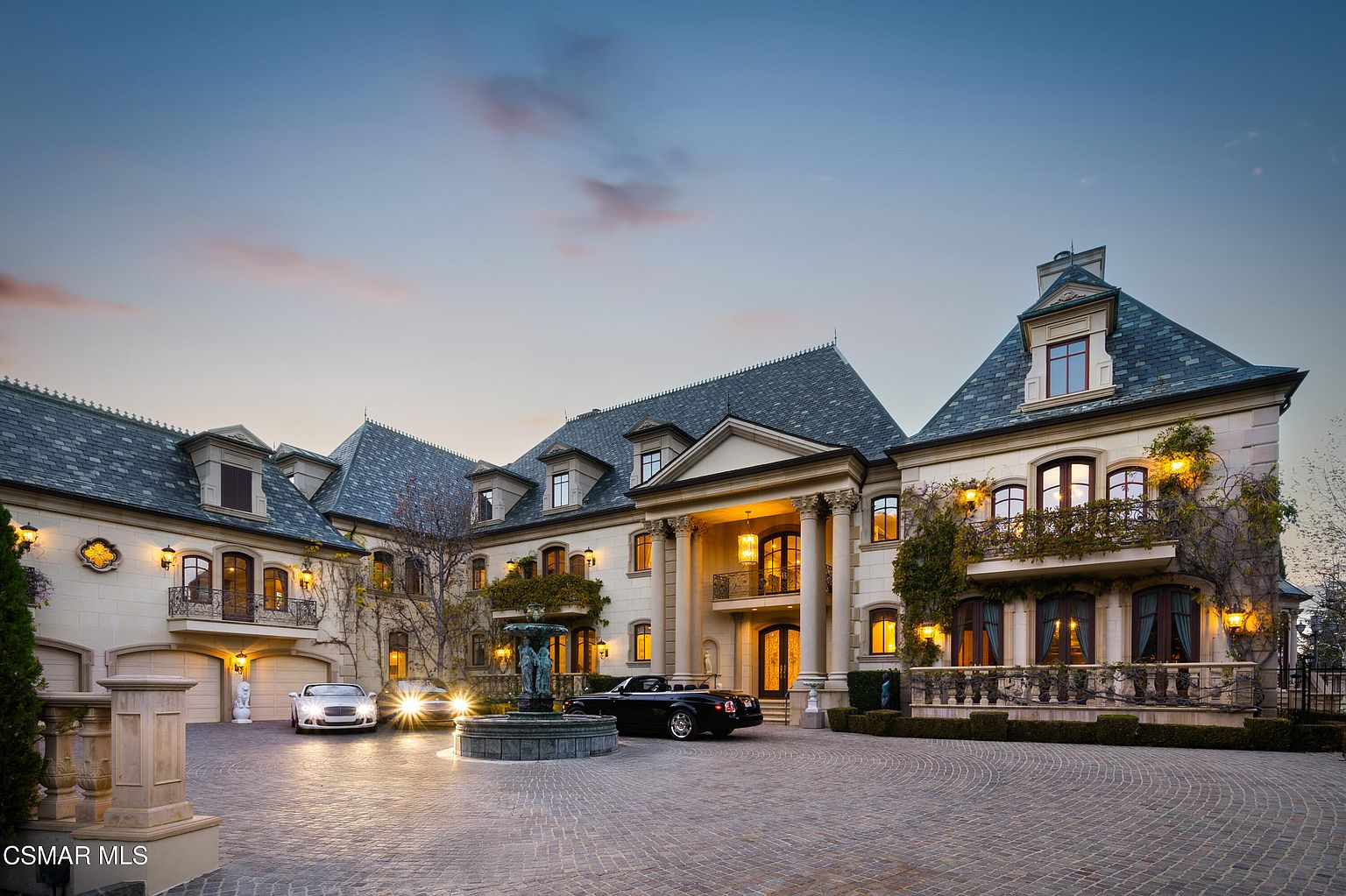 This grand, French-inspired estate showcases an impressive front facade with a symmetrical design, stone masonry, and a prominent two-story portico supported by classical columns. A circular cobblestone driveway leads to a central fountain, flanked by luxury vehicles, while the exterior is accented with elegant wrought-iron balconies and lush climbing greenery. The warm glow of the evening lighting highlights the architectural sophistication and opulent scale of this luxury residence.