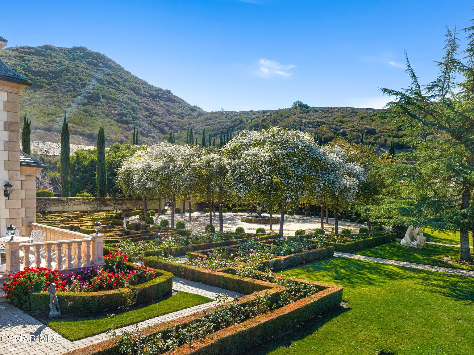 This stunning, meticulously manicured formal garden features intricate boxwood hedges, vibrant rose bushes, and a central gravel courtyard shaded by large, white-flowering trees. The scene is framed by a stone terrace in the foreground and a dramatic, sun-drenched hillside backdrop, creating an atmosphere of refined elegance and tranquility. The elevated perspective captures the expansive layout and the harmonious blend of structured landscaping with the natural beauty of the surrounding terrain.