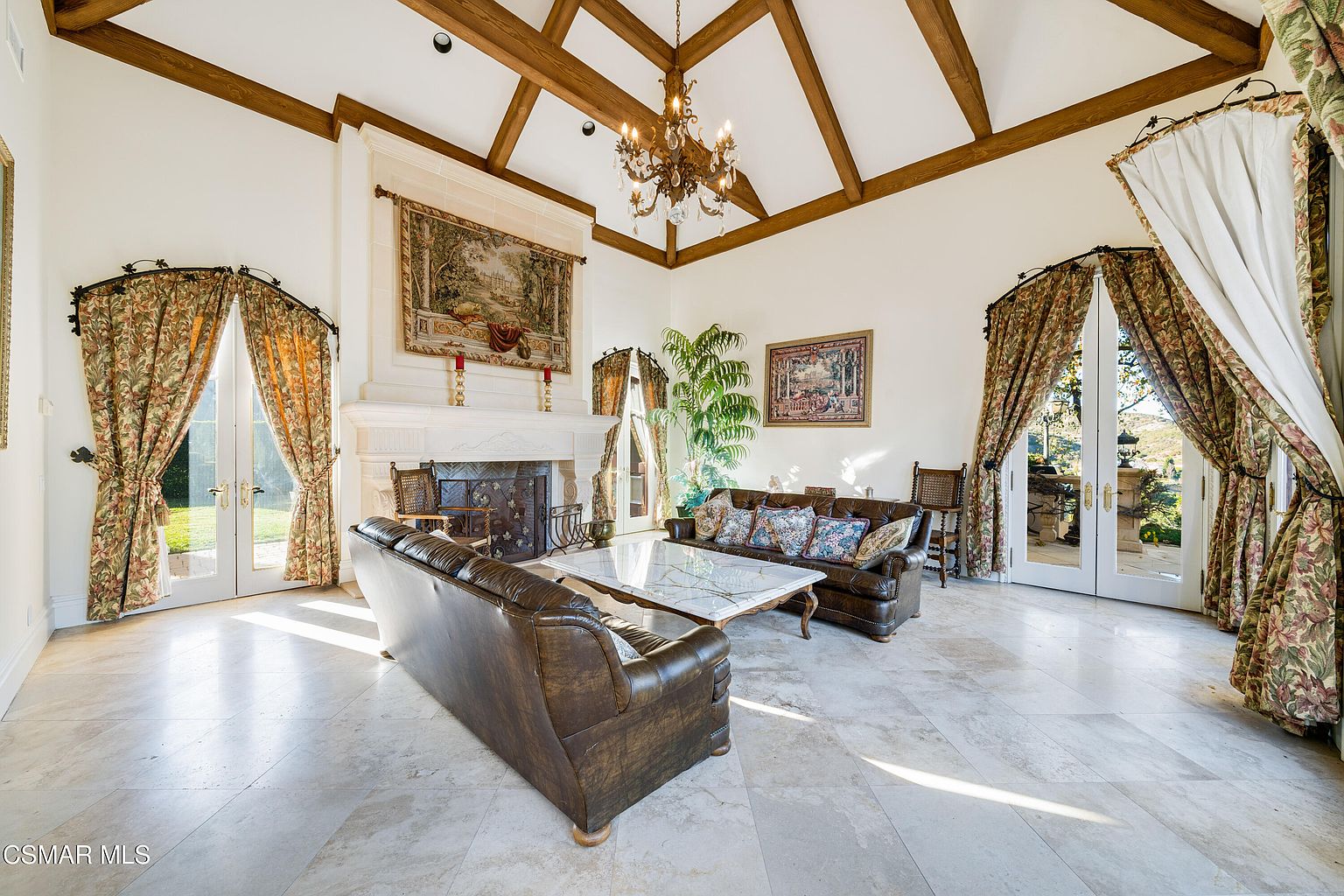 This grand living room features soaring vaulted ceilings with exposed wooden beams and a large, ornate chandelier as the centerpiece. The space is anchored by a stately stone fireplace adorned with a decorative tapestry, flanked by two sets of French doors draped in elegant floral curtains that provide abundant natural light. The room is furnished with plush leather sofas and a marble-topped coffee table, creating a sophisticated and luxurious atmosphere.