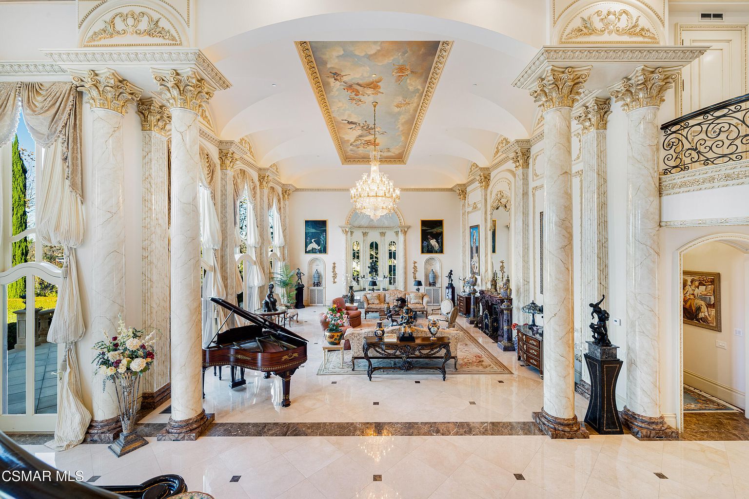 This grand, opulent living room features soaring ceilings with a detailed mural, massive marble columns, and a crystal chandelier that serves as a dramatic focal point. The space is furnished with elegant, traditional seating, a grand piano, and intricate decorative accents, all set against a backdrop of polished marble floors. The perspective is captured from an elevated vantage point, emphasizing the room's vast scale and luxurious, palatial aesthetic.