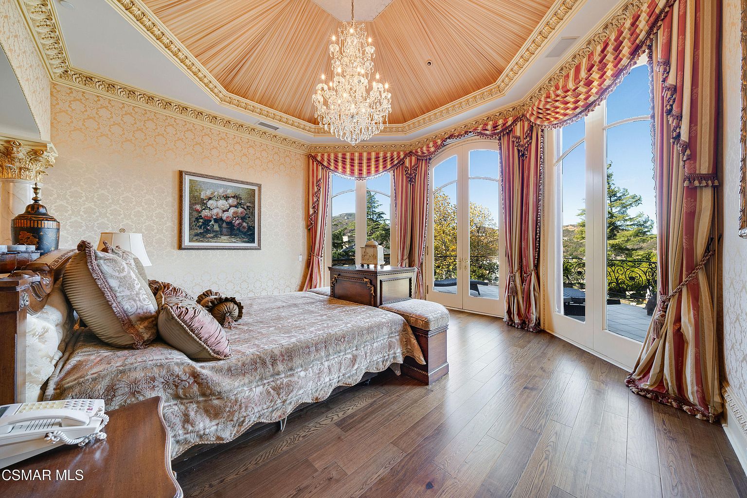 This opulent primary bedroom features a dramatic vaulted ceiling with a central crystal chandelier and elegant, patterned wallpaper. The room is anchored by a large bed with ornate bedding, positioned to face floor-to-ceiling arched glass doors that open onto a private balcony with scenic views. The combination of rich wood flooring, classic drapery, and sophisticated architectural details creates a luxurious and serene atmosphere.