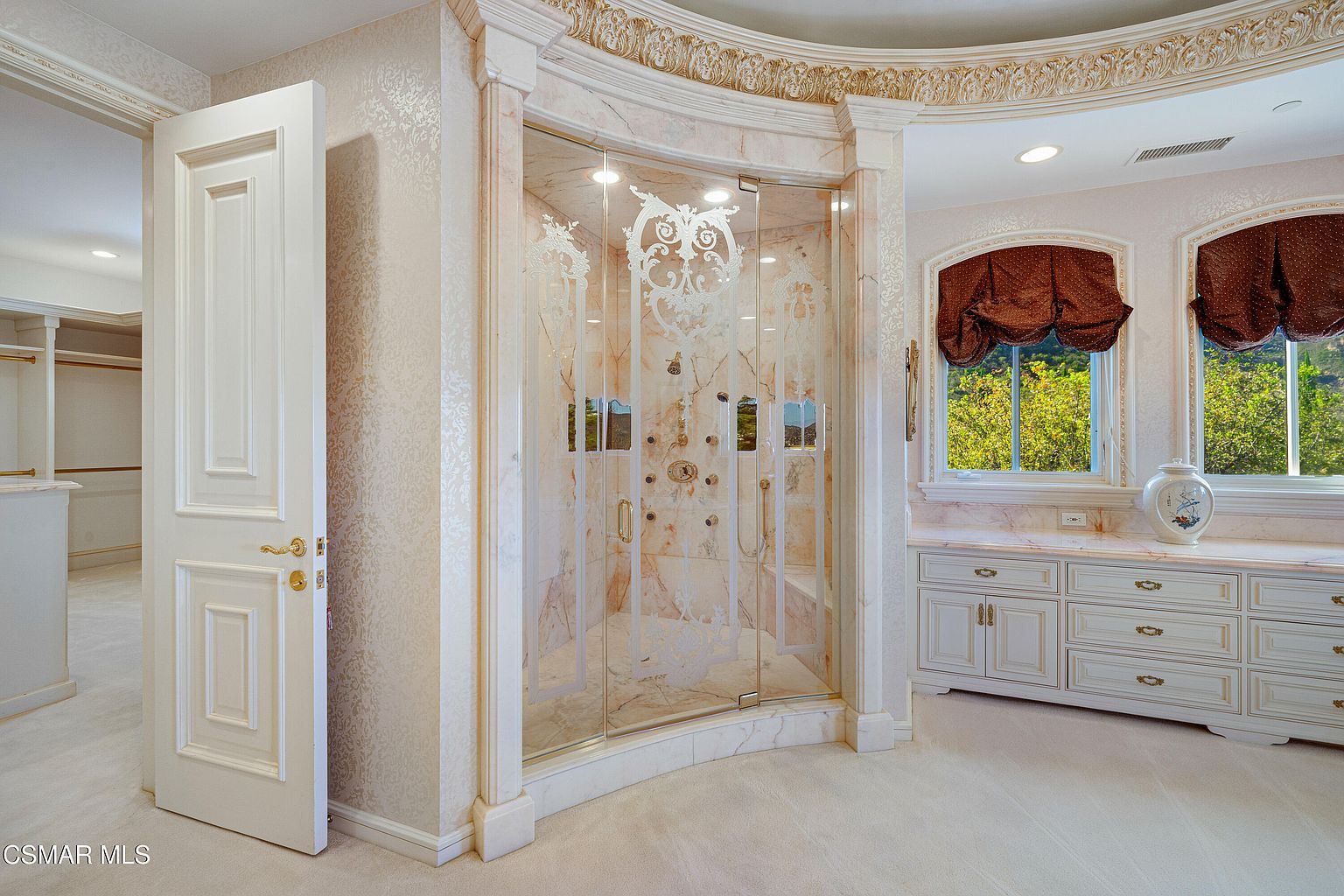 This luxurious primary bathroom features a grand, curved glass-enclosed shower with intricate etched detailing and marble tile work. The space is complemented by custom cabinetry with a marble countertop, elegant wallpaper, and a soft, neutral color palette that exudes opulence. The perspective captures the sophisticated layout, highlighting the seamless transition between the vanity area and the walk-in shower.