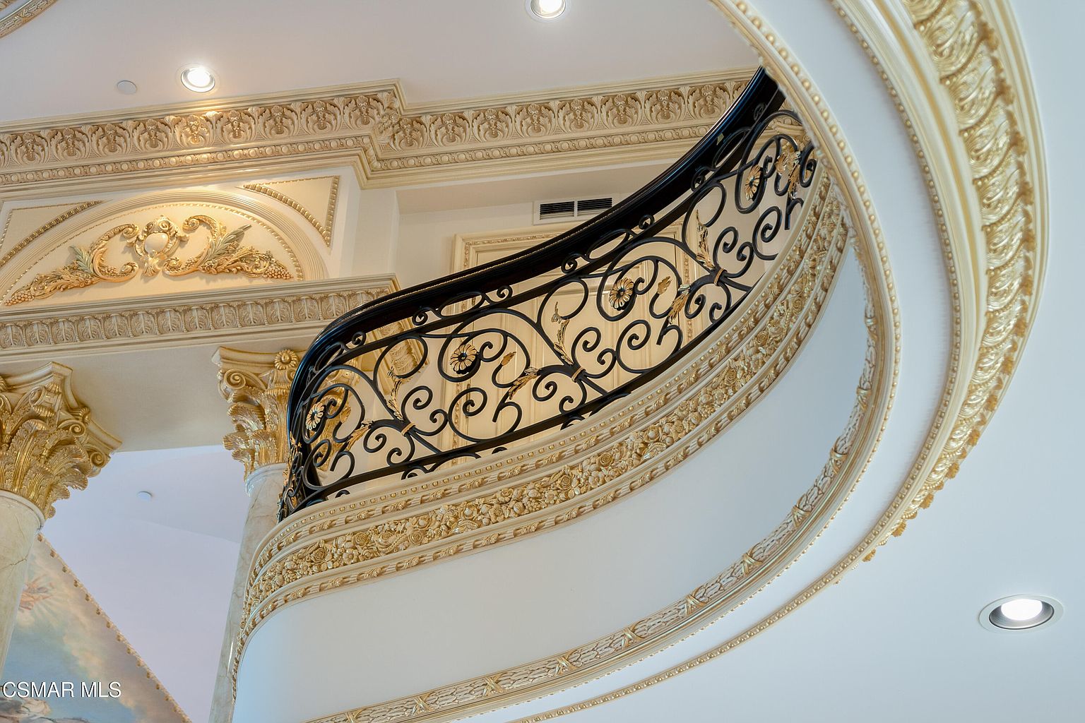This image captures a grand, ornate staircase featuring an intricate black wrought-iron railing with elegant scrollwork. The architecture is defined by opulent gold-leaf detailing, classical molding, and a sweeping curved design that exudes luxury and sophistication. The perspective is angled upward, emphasizing the height and the elaborate craftsmanship of the ceiling and wall trim.