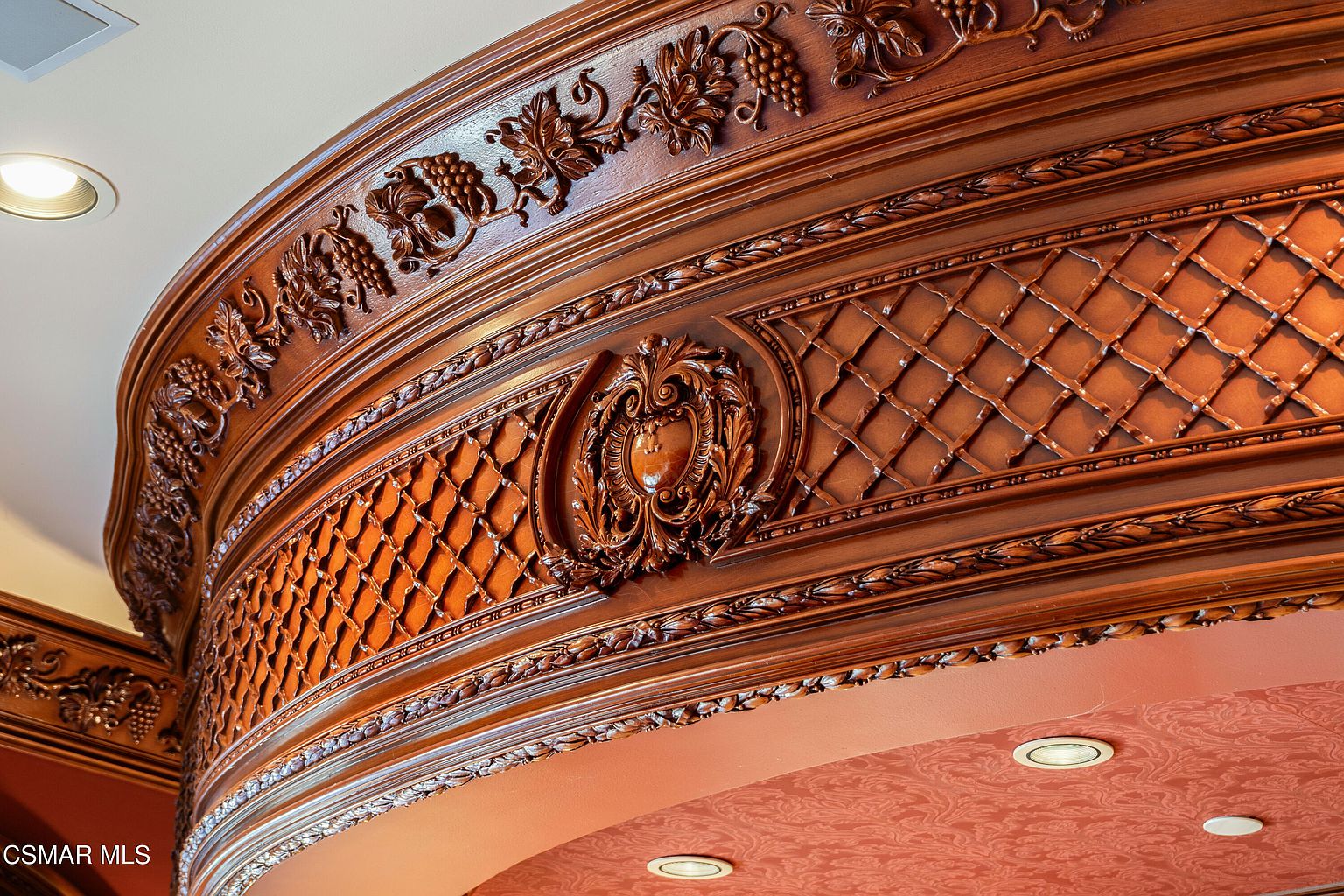This image captures a highly ornate, custom-carved wooden architectural detail, likely part of a grand ceiling cove or crown molding. The craftsmanship features intricate grape vine motifs and a central shield emblem, set against a lattice-patterned background that adds depth and texture. The warm, rich wood tones and sophisticated design suggest a luxurious, traditional, or Mediterranean-inspired interior aesthetic.
