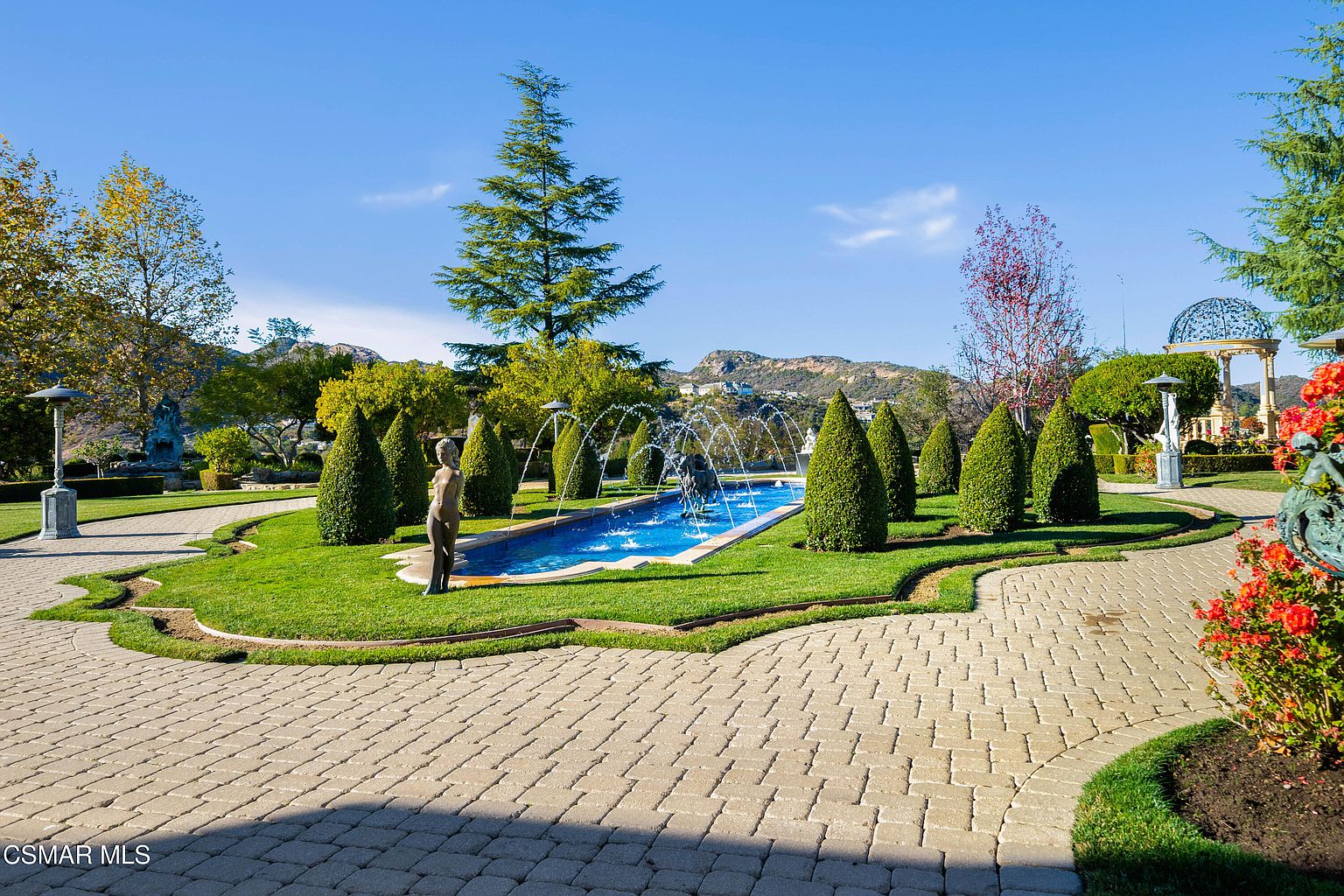 This expansive, meticulously landscaped garden features a long, rectangular reflecting pool with integrated fountains as its centerpiece. The scene is framed by manicured conical hedges, classical statues, and a stone-paved walkway, all set against a backdrop of rolling hills and a clear blue sky. A decorative gazebo stands in the distance, contributing to the estate's elegant, formal, and serene atmosphere.