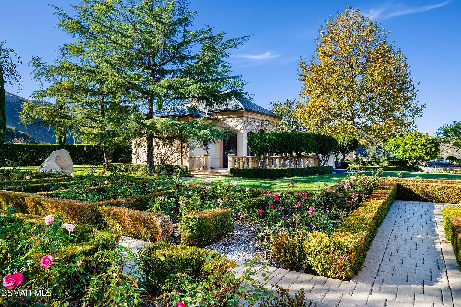 This picturesque garden scene features a meticulously manicured rose garden with geometric boxwood hedges and a stone paver walkway leading toward a charming, stone-clad garden pavilion. The property is framed by mature, lush trees and vibrant flowers, creating a serene and elegant atmosphere. The bright, clear sky and well-maintained landscaping suggest a high-end, tranquil estate setting.