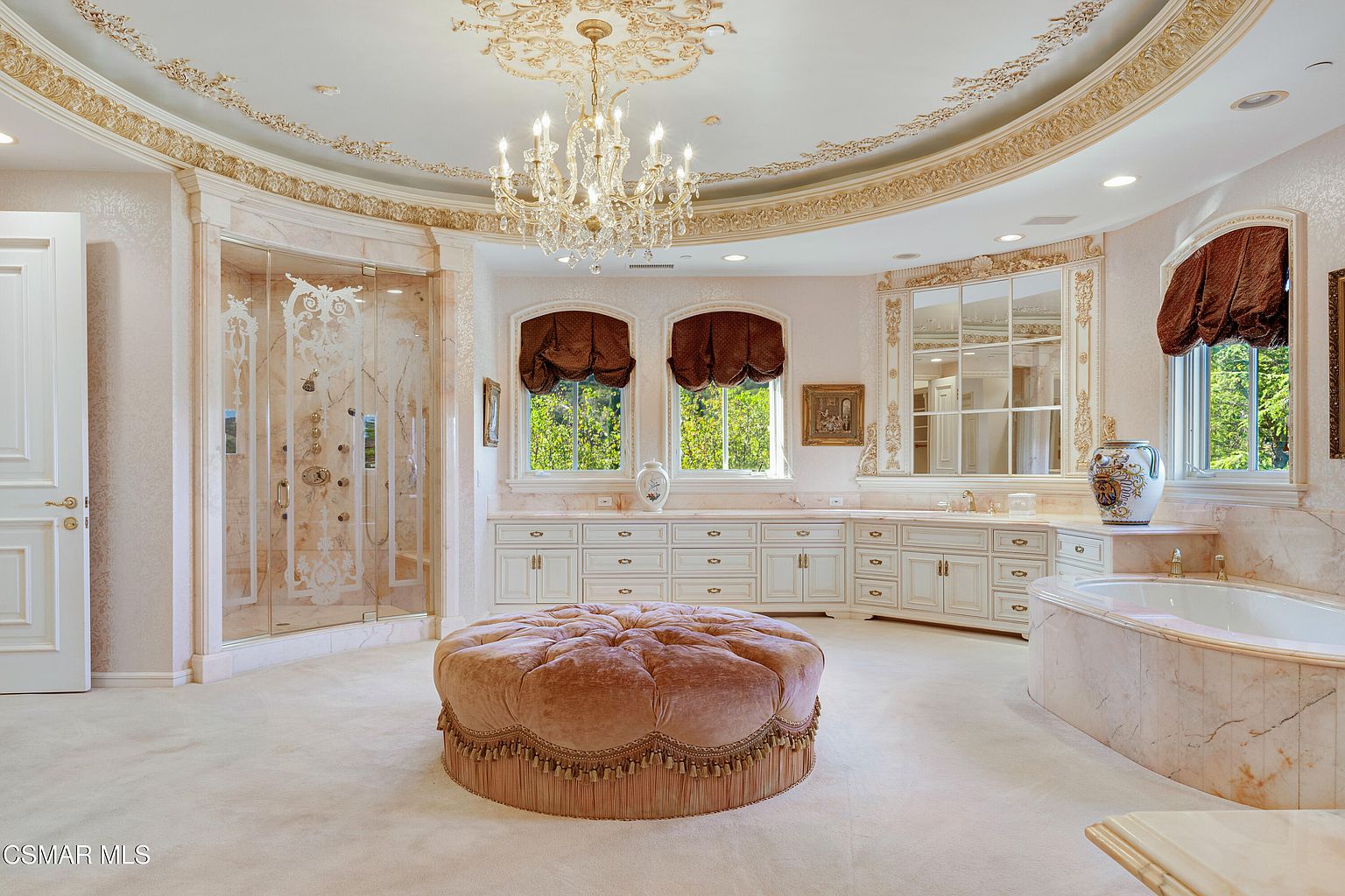 This opulent primary bathroom features a grand, circular layout with a central tufted velvet ottoman and a stunning crystal chandelier suspended from a decorative, ornate ceiling medallion. The space is finished in luxurious marble, including a large walk-in glass shower and a built-in soaking tub, complemented by custom cabinetry and elegant window treatments. The overall aesthetic is one of classic, high-end luxury and sophisticated design.