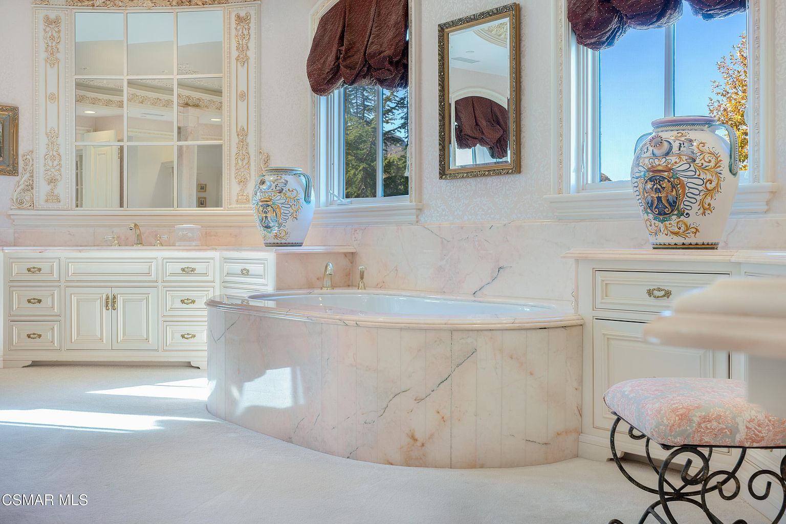 This luxurious primary bathroom features a grand, marble-encased soaking tub as its centerpiece, flanked by ornate, hand-painted ceramic vases. The space is adorned with elegant white cabinetry, intricate gold-leaf detailing on the mirrors, and plush carpeting, creating a sophisticated and opulent atmosphere. The perspective is eye-level, capturing the refined craftsmanship and classic, high-end design elements of the room.