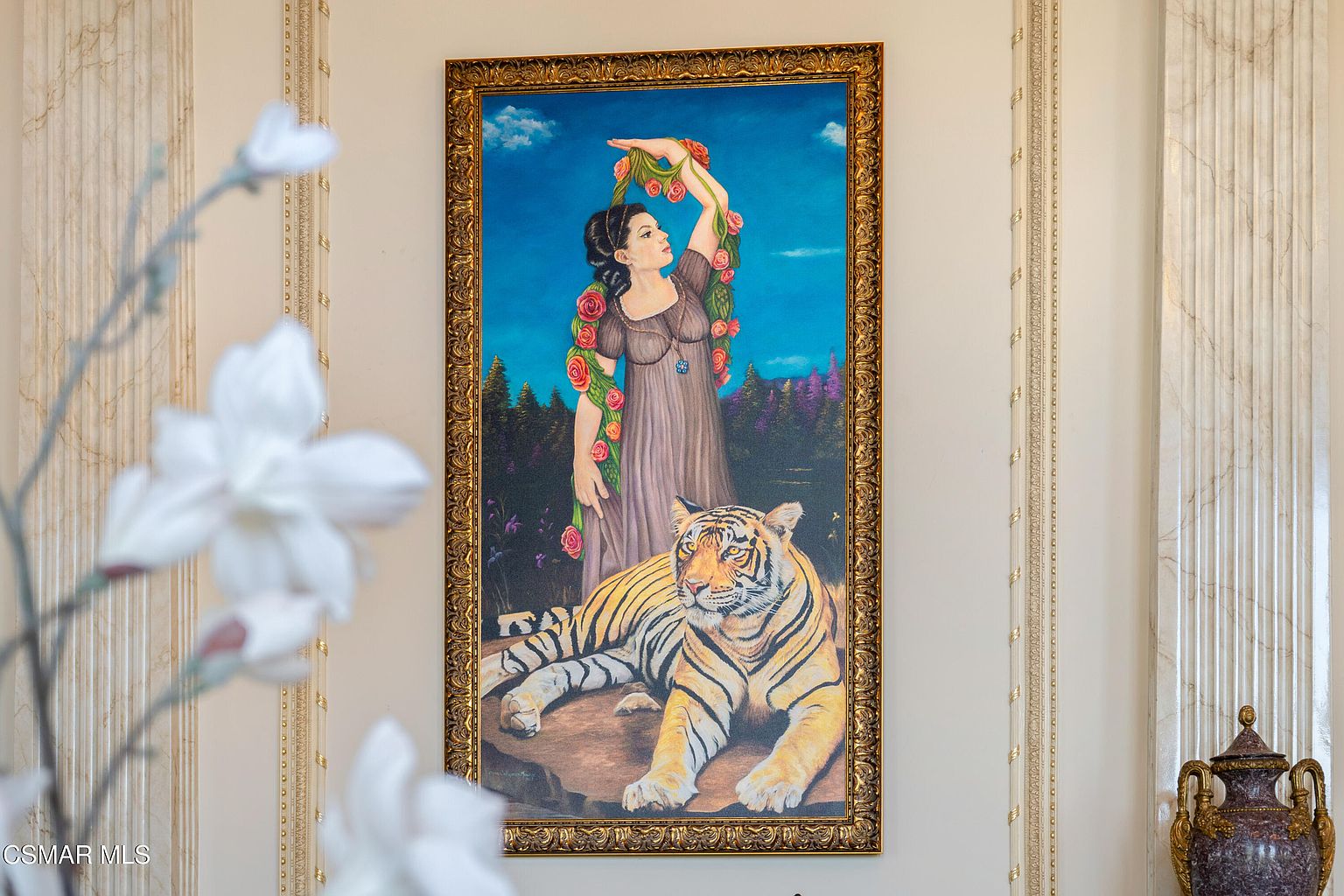 This image captures a sophisticated interior detail featuring a large, ornate framed painting of a woman with a tiger, mounted on a wall with elegant, fluted architectural columns. The foreground is softly blurred with white floral elements, creating a sense of depth and luxury. The overall aesthetic is opulent and classical, highlighting the refined decor of the home.