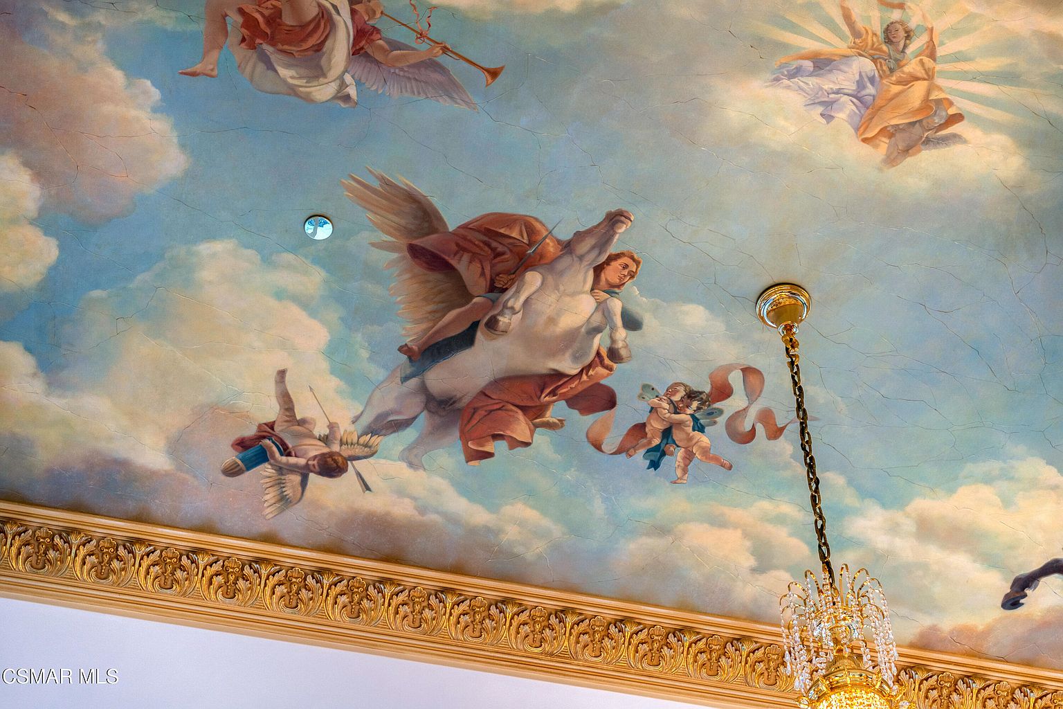 This image captures a dramatic, ornate ceiling mural featuring a classical mythological scene with a winged horse, cherubs, and celestial figures set against a painted sky. The artwork is framed by an elaborate, gilded crown molding and is complemented by a hanging crystal chandelier that adds a touch of luxury. The perspective is looking directly upward, emphasizing the grandeur and artistic detail of the ceiling design.