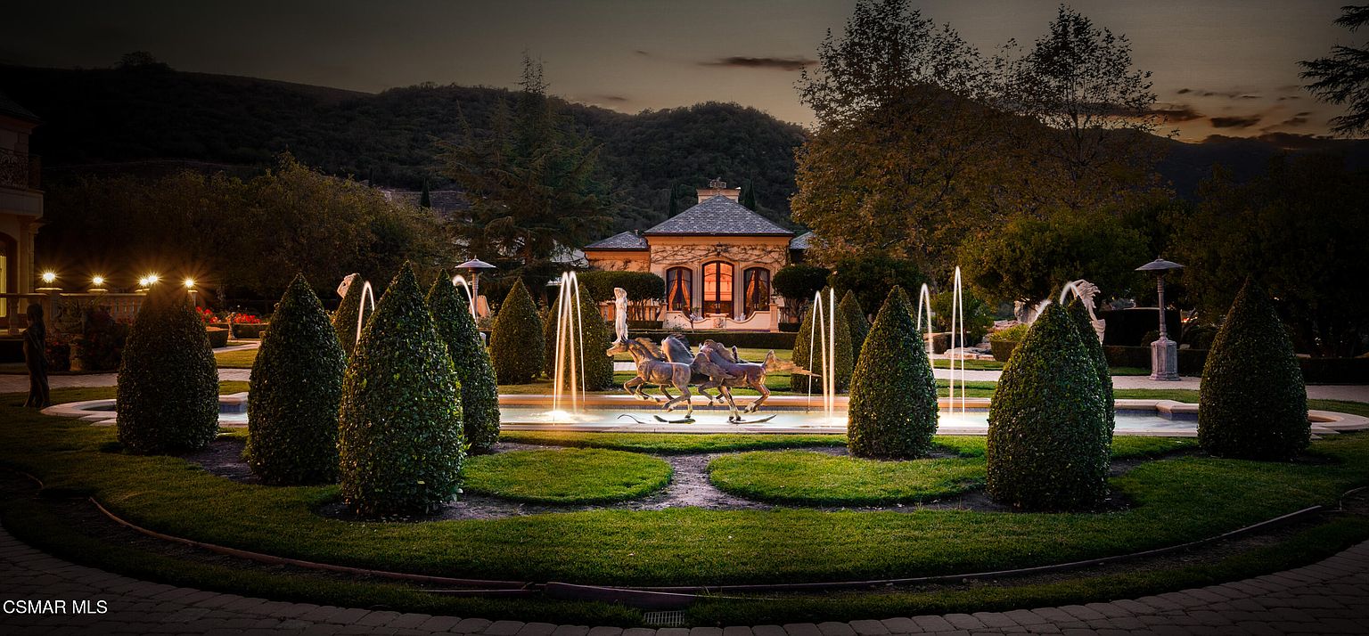 This stunning, professionally landscaped garden features a central fountain with a dramatic bronze horse sculpture, illuminated by soft evening lighting. The scene is framed by perfectly manicured conical hedges and lush green lawns, leading the eye toward a sophisticated, Mediterranean-style estate building in the background. The twilight setting creates an elegant, serene, and luxurious atmosphere, highlighting the property's high-end curb appeal.