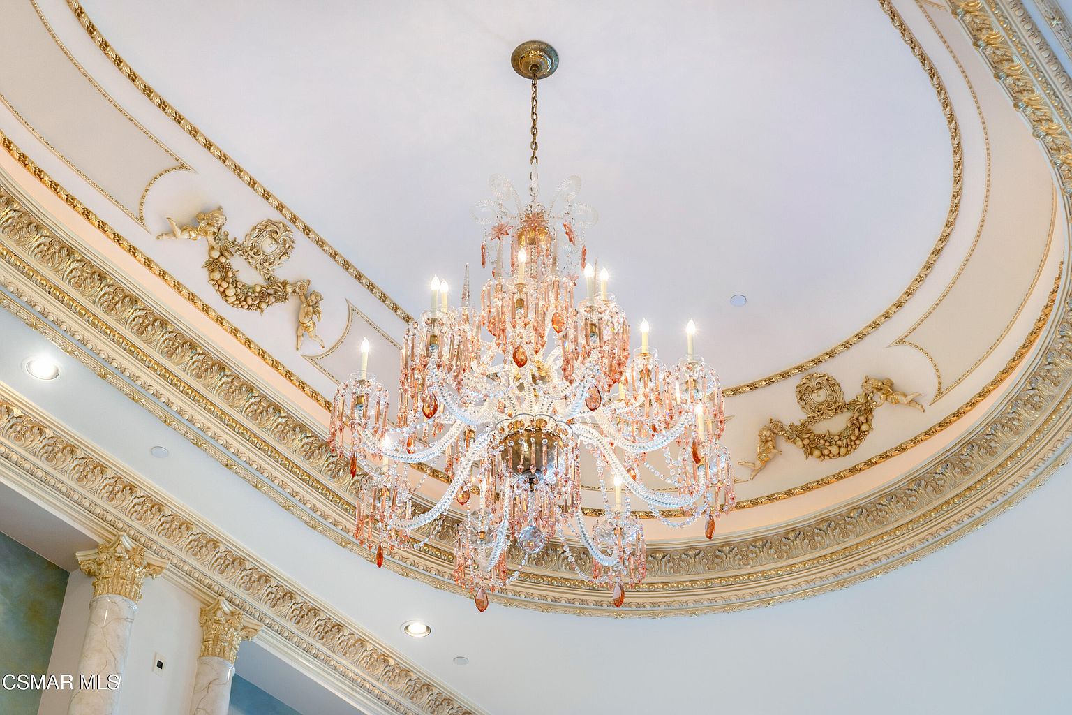 This image captures an opulent, ornate ceiling featuring a large, multi-tiered crystal chandelier as the centerpiece. The ceiling is adorned with intricate gold-leaf molding, decorative relief carvings of cherubs and floral garlands, and a recessed circular design that adds depth and grandeur. The perspective is looking directly upward, emphasizing the luxurious craftsmanship and the dramatic scale of the lighting fixture.