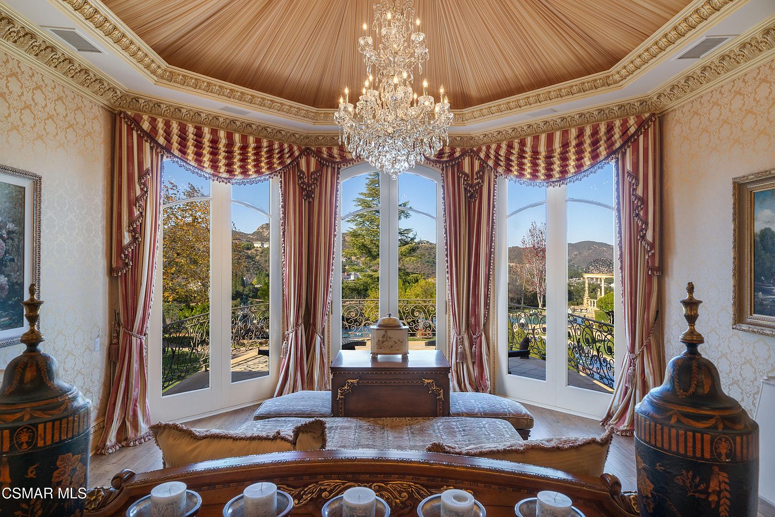 This elegant, formal living space features a dramatic domed ceiling with a grand crystal chandelier and ornate gold-trimmed crown molding. Large, floor-to-ceiling arched windows draped in luxurious, multi-layered fabric curtains frame a picturesque view of the rolling hills and outdoor balcony. The room is anchored by a central wooden console table and plush seating, creating a sophisticated and opulent atmosphere.