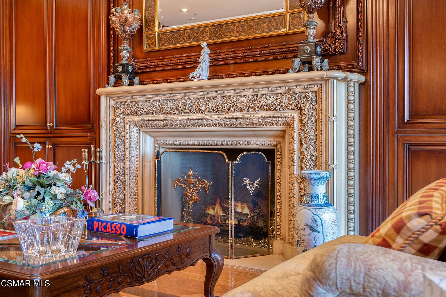 This elegant living room features a grand, ornate fireplace with intricate floral carvings and a warm, glowing fire behind a decorative metal screen. The space is framed by rich, dark wood paneling and a large, gilded mirror above the mantel, creating a sophisticated and traditional atmosphere. A coffee table in the foreground holds a book on Picasso, while a plush, patterned armchair and a floral arrangement add comfort and color to the refined setting.