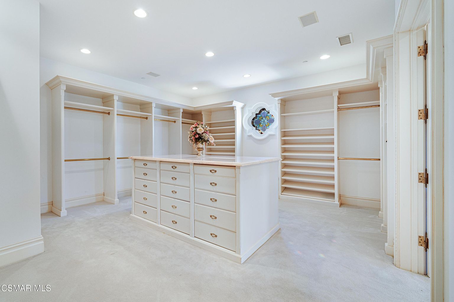 This spacious, high-end walk-in closet features custom built-in cabinetry with ample hanging space, open shelving, and a central island topped with marble. The room is illuminated by recessed lighting and accented by a decorative stained-glass wall feature, creating a luxurious and organized atmosphere. The neutral color palette and plush carpeting enhance the sophisticated, clean aesthetic of this primary suite amenity.