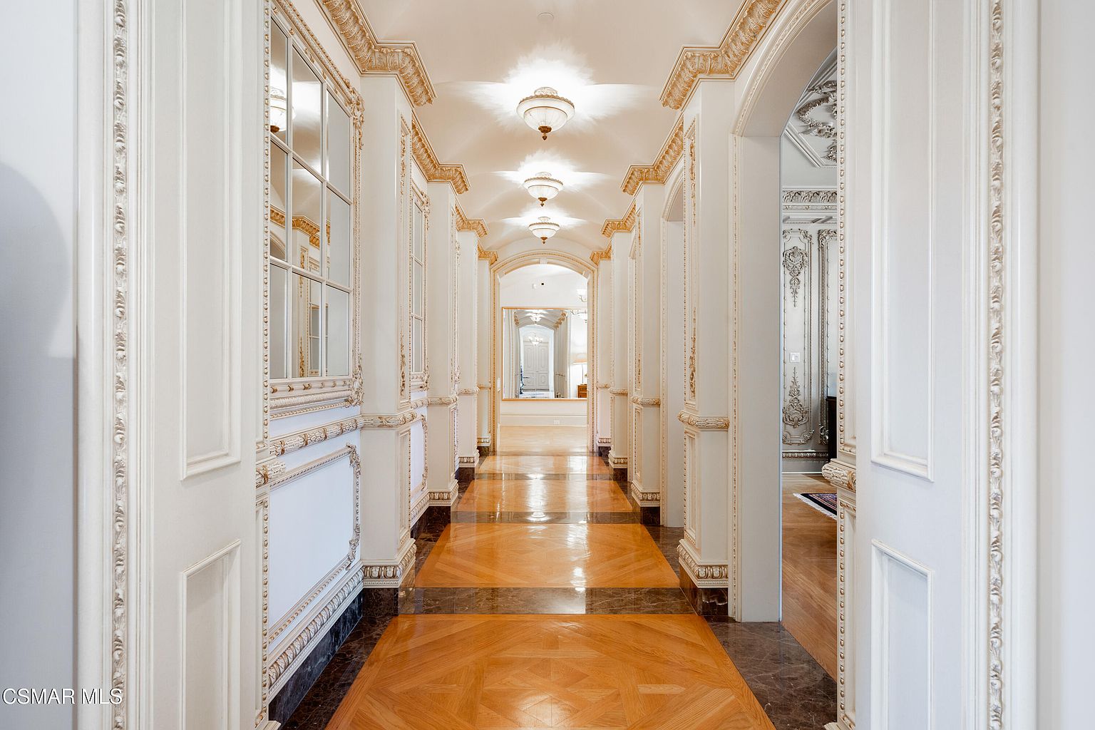 This opulent, grand hallway features intricate gold-leaf crown molding, ornate wall paneling, and a sophisticated mix of polished hardwood and dark marble flooring. The perspective is a centered, symmetrical shot looking down the long corridor, which is illuminated by a series of elegant, glowing ceiling-mounted light fixtures. The overall aesthetic is one of classical luxury and high-end architectural detail, creating a sense of regal scale and refinement.