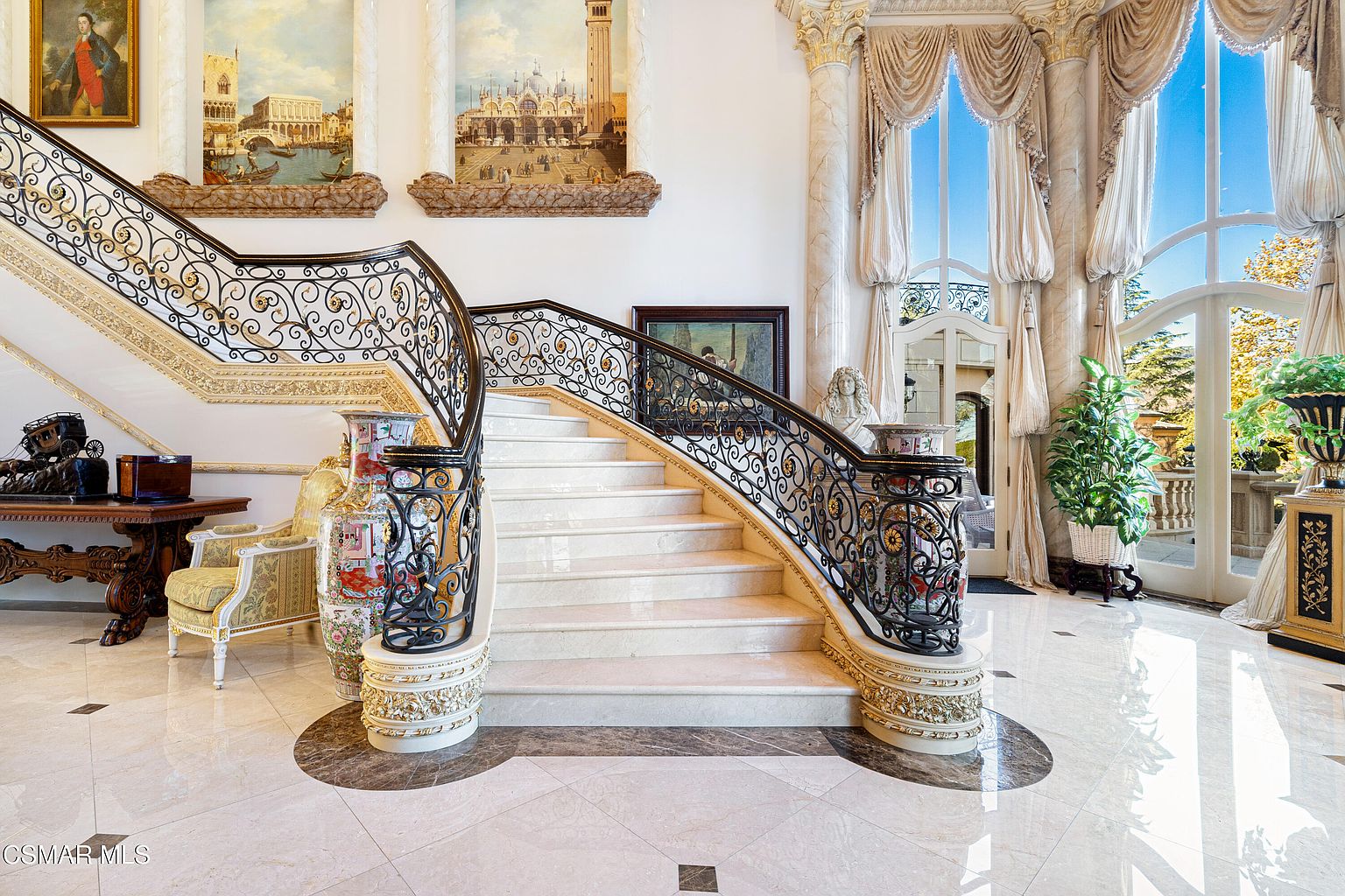 This grand foyer features a sweeping, ornate marble staircase with intricate wrought-iron railings and gold accents, serving as the dramatic centerpiece of the space. The room is adorned with classical artwork, large decorative vases, and towering windows draped in elegant fabric, creating an atmosphere of opulent, old-world luxury. The perspective is centered and symmetrical, emphasizing the height of the ceilings and the high-end architectural craftsmanship.