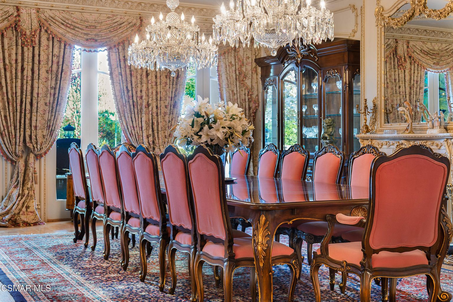 This opulent formal dining room features a long, polished wooden table surrounded by elegant chairs with pink upholstered seats and backs. The space is illuminated by two grand crystal chandeliers hanging from the ceiling, while heavy, ornate floral-patterned drapes frame the large windows. A dark wood china cabinet stands against the wall, and a rich, traditional area rug anchors the room, creating a sophisticated and luxurious atmosphere.