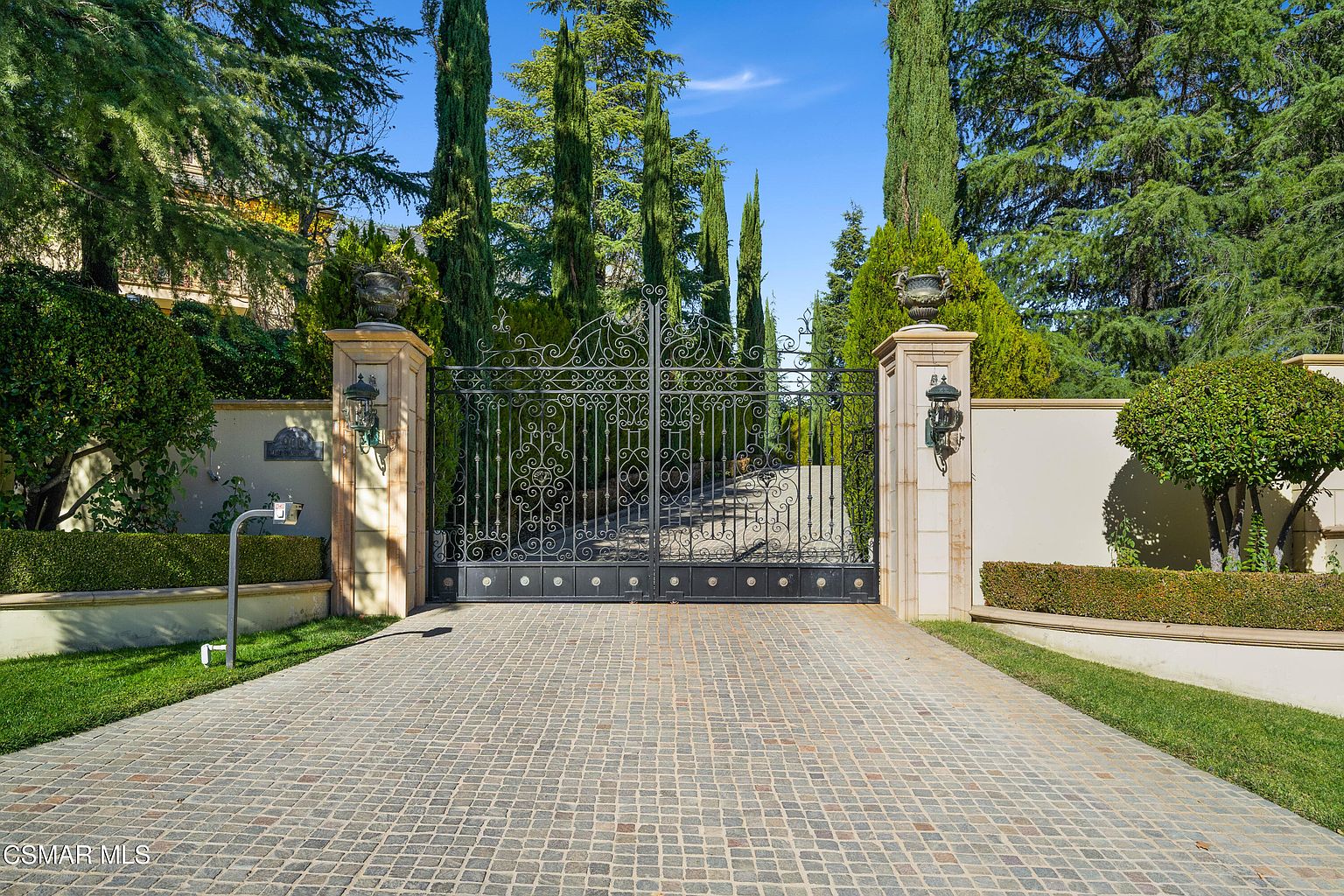 This image showcases a grand, gated entrance to a luxury estate, featuring ornate black wrought-iron gates flanked by stone pillars topped with decorative urns. A meticulously paved cobblestone driveway leads up to the gates, framed by lush green hedges, manicured lawns, and towering cypress trees. The scene conveys an atmosphere of exclusivity, privacy, and high-end architectural elegance.