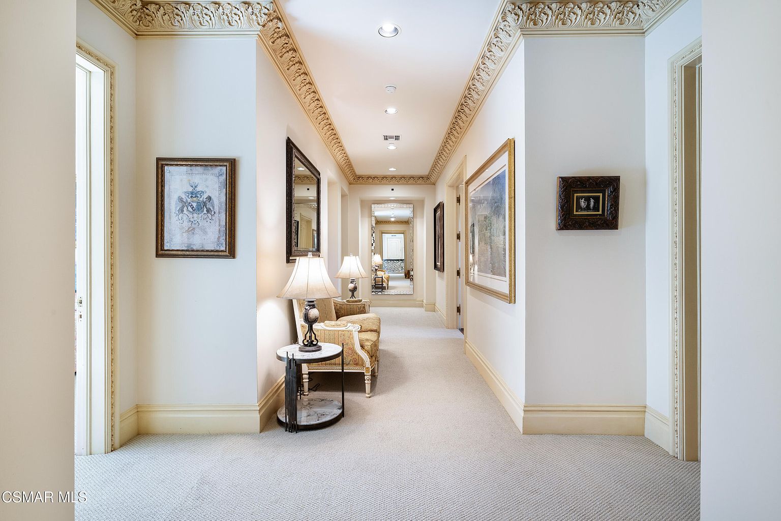 This elegant, long hallway features ornate crown molding and a neutral carpet, creating a sophisticated and classic atmosphere. The space is furnished with a small side table, a lamp, and a chair, while framed artwork and a large mirror at the end of the hall add depth and visual interest. The perspective is a centered, eye-level shot that draws the viewer down the length of the corridor, emphasizing the home's spacious and refined layout.