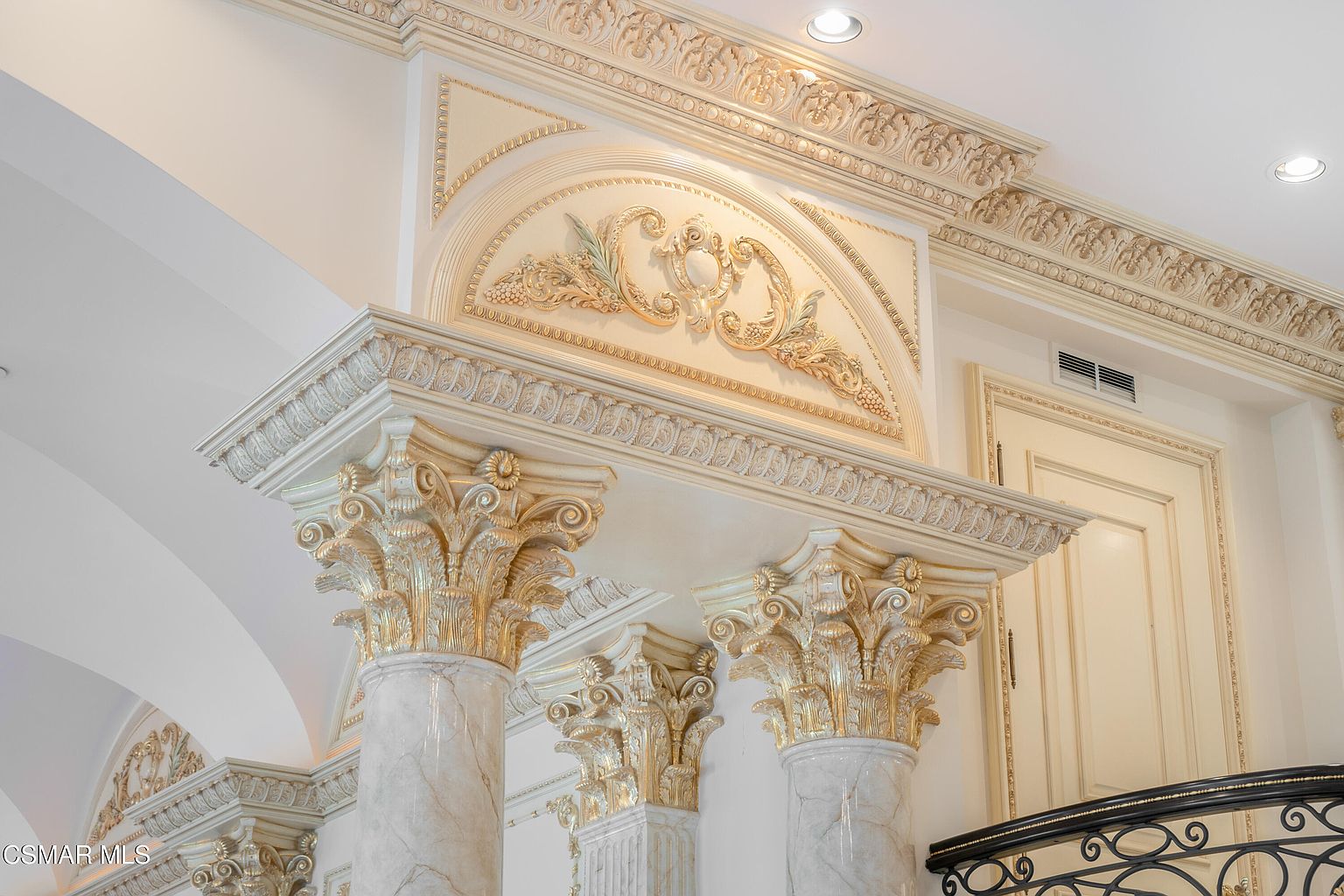 This image captures a highly ornate, neoclassical architectural detail featuring marble columns topped with elaborate, gold-leaf accented Corinthian capitals. Above the columns, a decorative pediment showcases intricate relief carvings of foliage and scrollwork, set against a cream-colored wall with extensive crown molding. The perspective is a low-angle, upward-looking shot that emphasizes the grandeur and opulent craftsmanship of the home's interior design.
