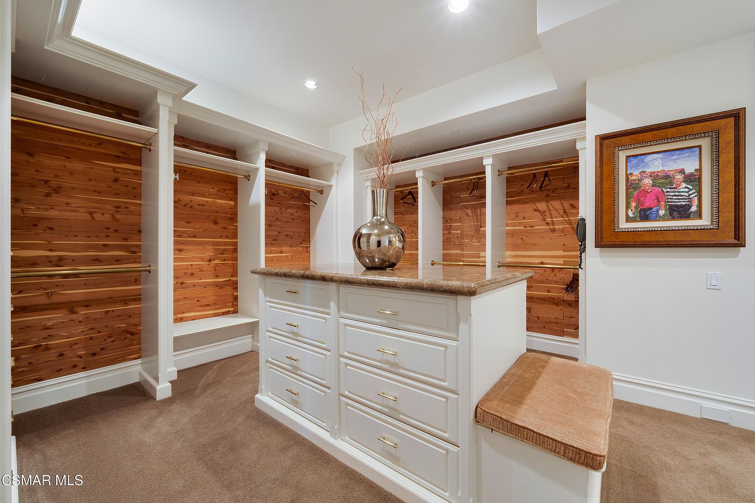 This spacious walk-in closet features custom white cabinetry with gold hardware, complemented by warm cedar wood paneling on the back walls. A central island with a stone countertop provides additional storage and a surface for display, while a built-in bench offers a convenient seating area. The room is finished with plush carpeting and recessed lighting, creating a luxurious and organized atmosphere.