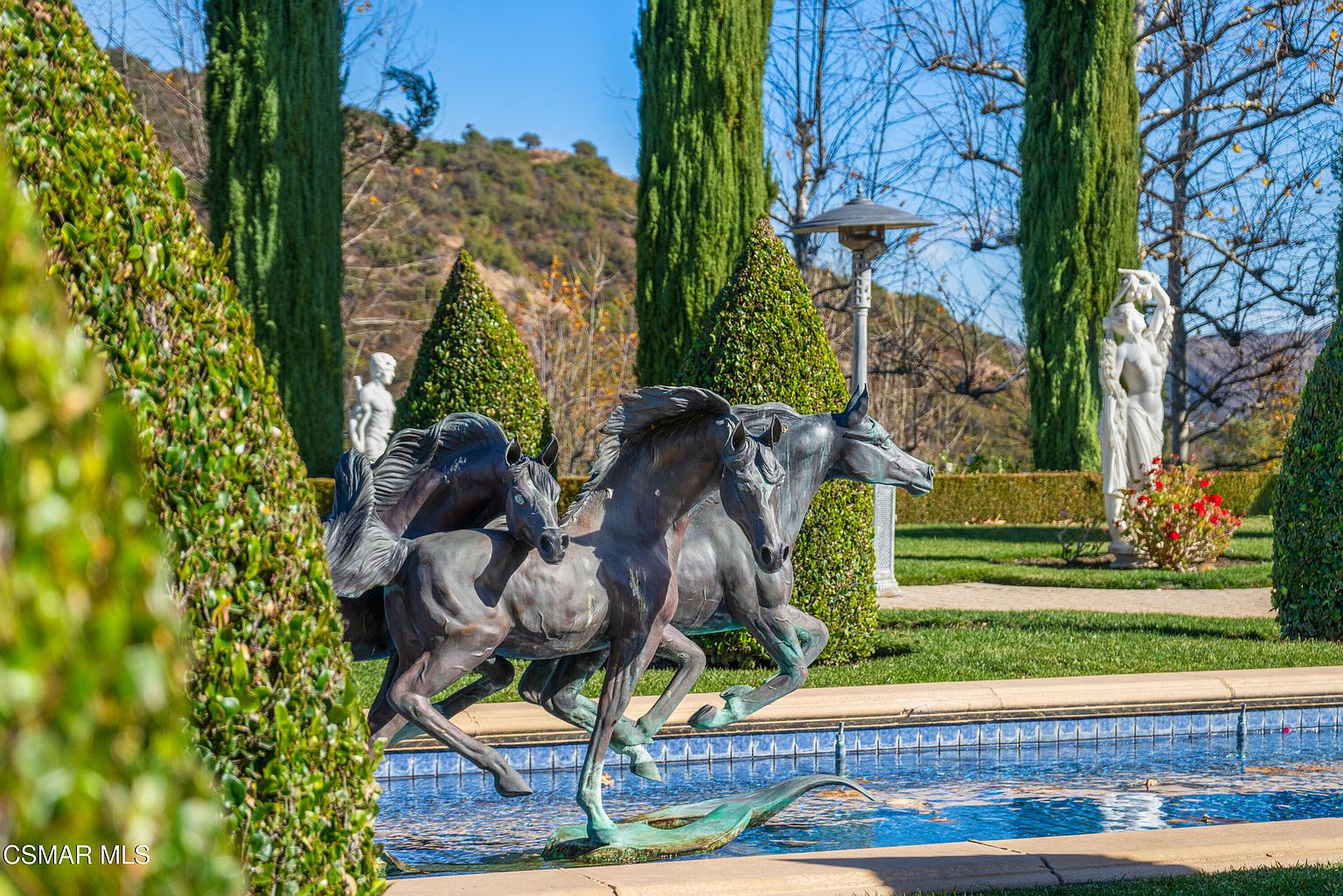 This elegant garden scene features a prominent bronze horse sculpture fountain as the central focal point, set against a backdrop of manicured topiary trees and a classical white statue. The composition captures a serene, formal landscape design with a blue-tiled water feature, suggesting a high-end, estate-like atmosphere. The perspective is eye-level, emphasizing the intricate details of the sculpture and the lush, well-maintained greenery of the surrounding grounds.