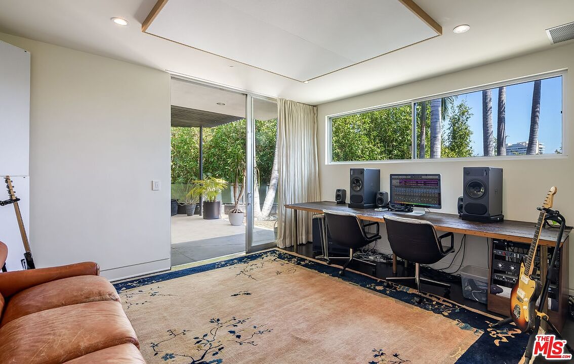 This interior shot showcases a modern office or study space with a large window providing natural light and views of greenery. The room features a long wooden desk with computer equipment and speakers, two black office chairs, and a tan rug with a blue floral pattern. A brown leather couch sits to the left, and a sliding glass door leads to an outdoor patio area.