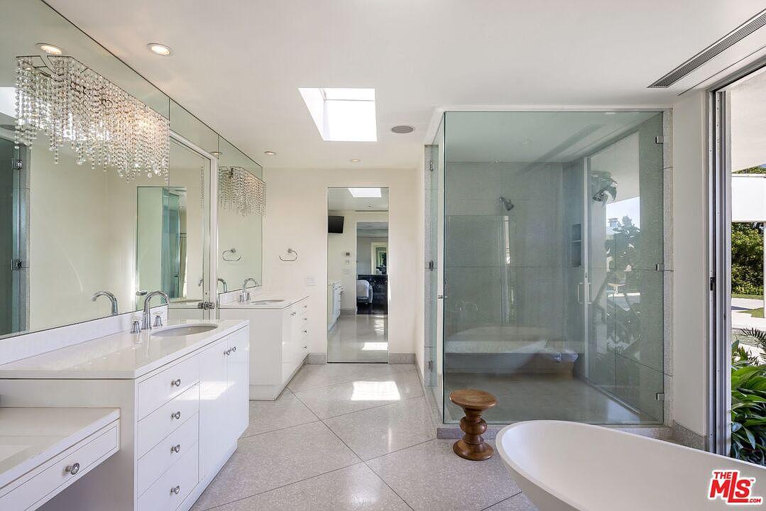 This is a bright and luxurious primary bathroom featuring a double vanity with white cabinetry and a large mirror adorned with a chandelier. A glass-enclosed shower and a freestanding bathtub are also visible, contributing to the bathroom's spa-like atmosphere. The flooring is a light-colored tile, and a skylight provides natural light.