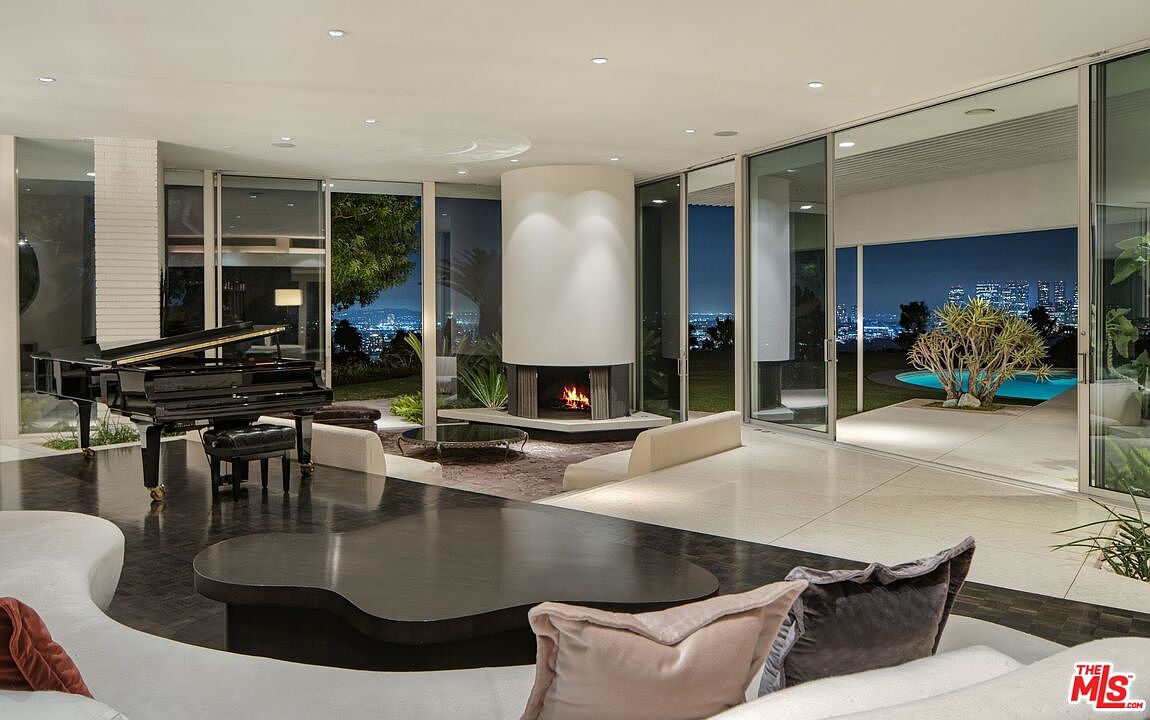 This is a luxurious living room featuring a grand piano, a modern fireplace, and floor-to-ceiling windows offering a stunning city view. The room is decorated with stylish furniture and has a sleek, open design that seamlessly connects to an outdoor pool area. The overall impression is one of elegance and sophistication.