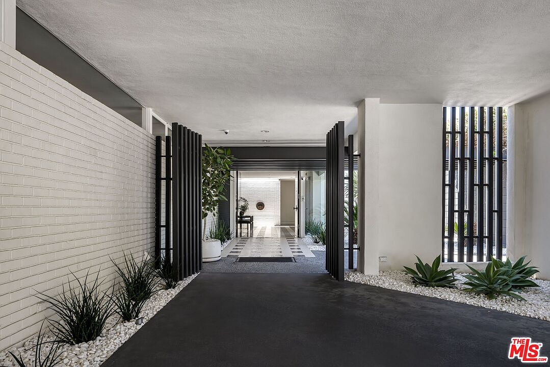 This image showcases a modern entryway with a striking design. A white brick wall contrasts with dark vertical slat screens, creating a visually appealing transition from the exterior to the interior. The pathway is lined with white gravel and minimalist plantings, leading to a bright, open doorway.