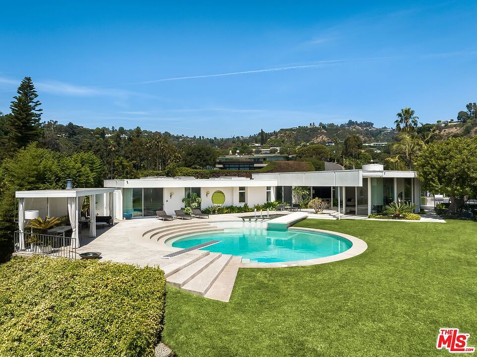 This image showcases a luxurious mid-century modern home with a stunning circular pool as its centerpiece. The pool is surrounded by a well-manicured lawn and features a unique stepped entrance. The house itself boasts clean lines, large windows, and a bright white exterior, blending seamlessly with the lush greenery and clear blue sky, creating an inviting and serene atmosphere.