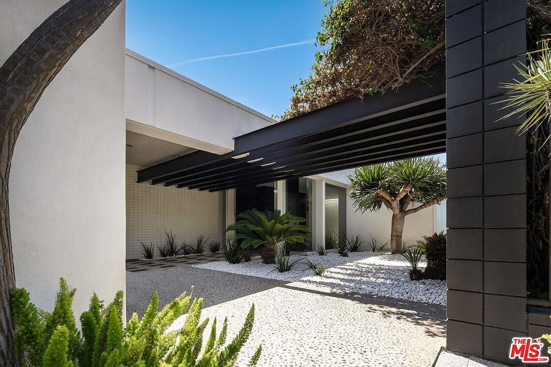 This image showcases the entryway of a modern home, featuring a striking black pergola structure and a minimalist garden design with white gravel and various plants. The architectural style is clean and contemporary, with a focus on geometric shapes and contrasting colors. The perspective is from the garden, looking towards the entrance, creating a welcoming and sophisticated impression.