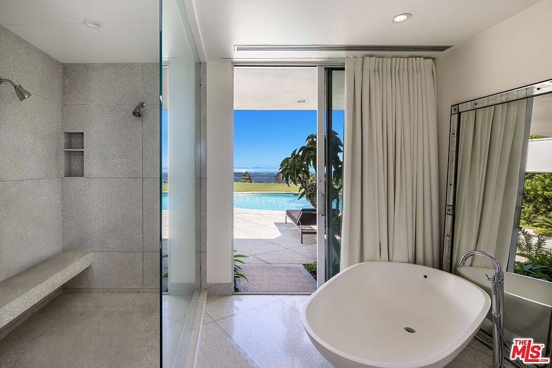 This is a luxurious primary bathroom featuring a modern freestanding bathtub, a spacious walk-in shower with a built-in bench, and a large mirror. The bathroom has an open design with a sliding glass door that leads to an outdoor pool area, offering a seamless indoor-outdoor living experience. The neutral color palette and clean lines create a serene and spa-like atmosphere.