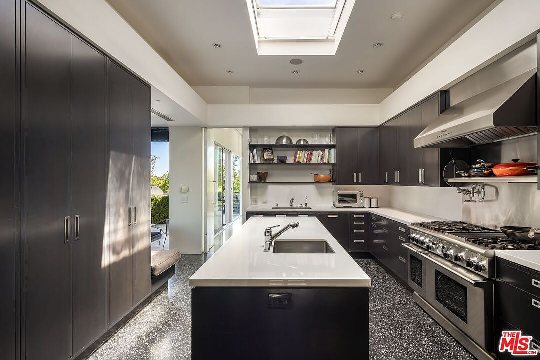 This is a modern kitchen with dark cabinetry, stainless steel appliances, and a large island with a white countertop. A skylight provides natural light, and open shelving displays decorative items. The kitchen has a sleek and minimalist design, with a focus on functionality and style, as seen from a wide angle.