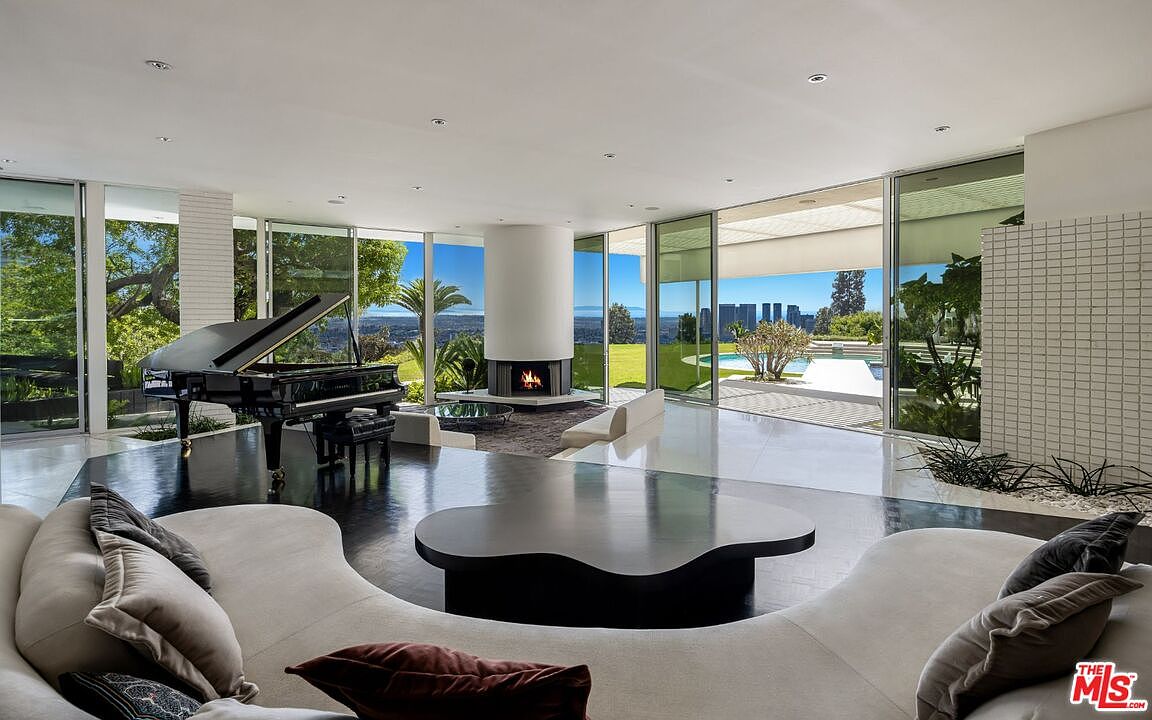 This is a spacious and luxurious living room featuring floor-to-ceiling windows that offer panoramic views of the city and landscape. The room includes a grand piano, a modern fireplace, and a unique, curved sofa with a dark, abstract-shaped coffee table. The polished floors and minimalist design contribute to an elegant and contemporary aesthetic.