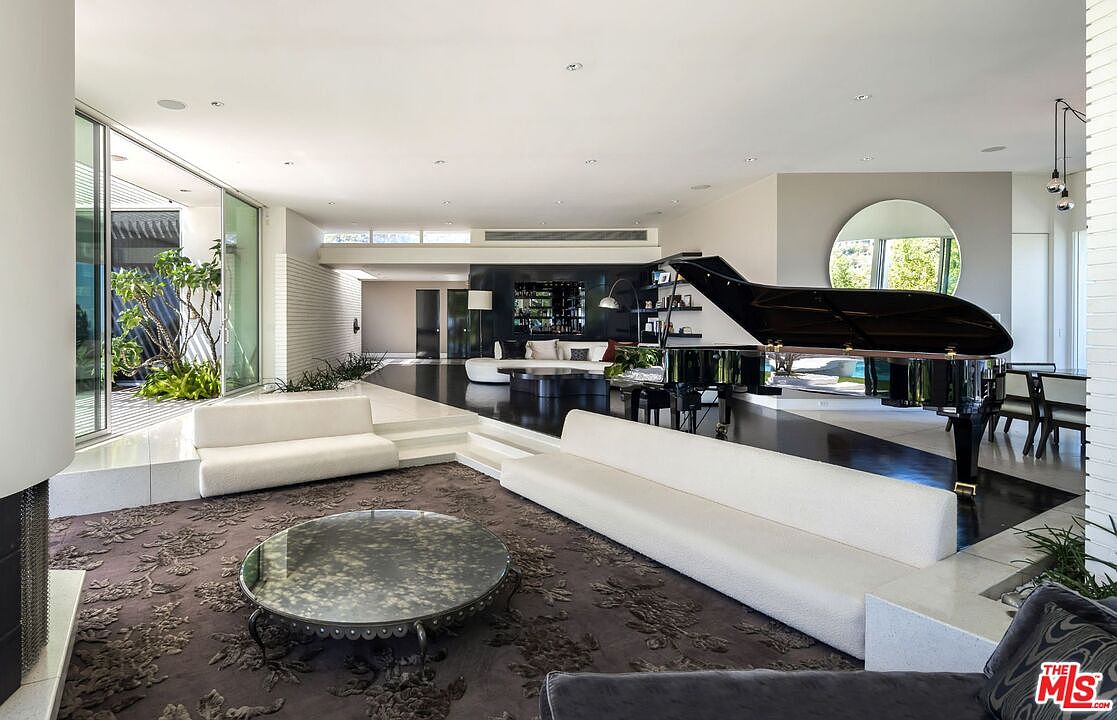 This is a spacious and luxurious living room featuring a grand piano, modern white sofas, and a unique round glass coffee table on a patterned rug. The room boasts a black and white tiled floor, a large round mirror, and an open layout with access to an outdoor courtyard, creating a sophisticated and airy atmosphere. The perspective is from a low angle, emphasizing the room's depth and grandeur.