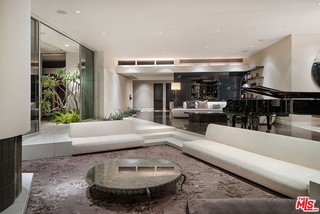 This is a spacious and modern living room featuring two white sofas, a round glass coffee table on a patterned rug, and a grand piano in the background. The room has a sunken area with dark flooring, a bar area, and a view of an indoor garden through large glass doors. The overall impression is luxurious and sophisticated.