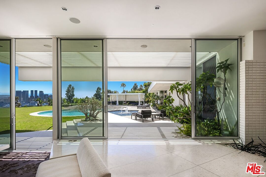 This image showcases a modern living room that seamlessly blends indoor and outdoor spaces through large sliding glass doors. The interior features sleek, light-colored tile flooring and minimalist furniture, while the exterior boasts a well-manicured lawn, a swimming pool, and a patio area with lounge seating. The open design and abundant natural light create an inviting and luxurious atmosphere.